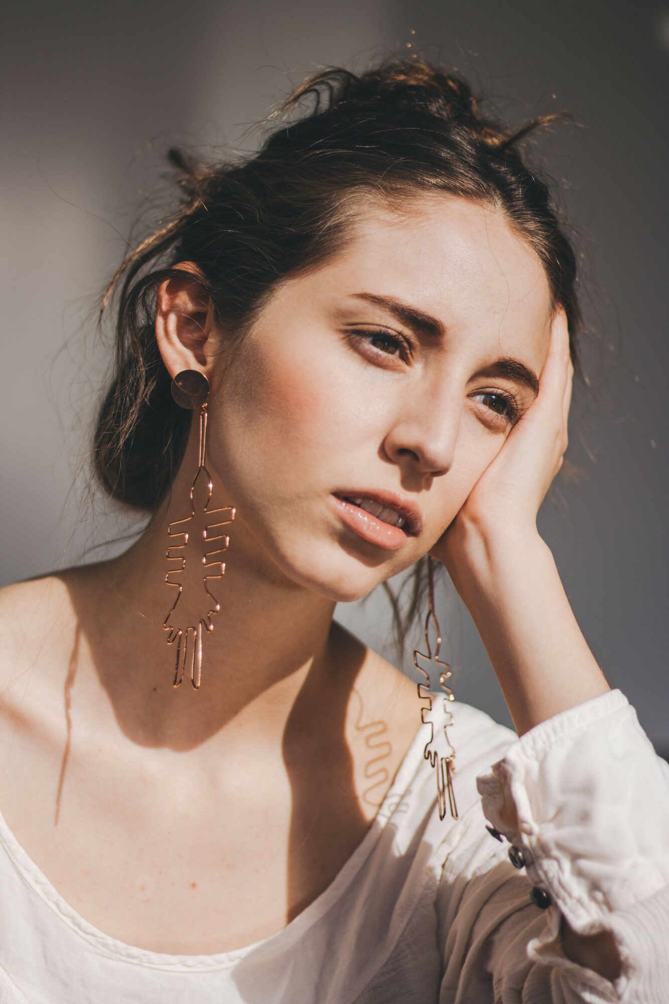 Young woman with dark hair, wearing large earrings, resting her head on her hand, looking away, sunlight on her face.
