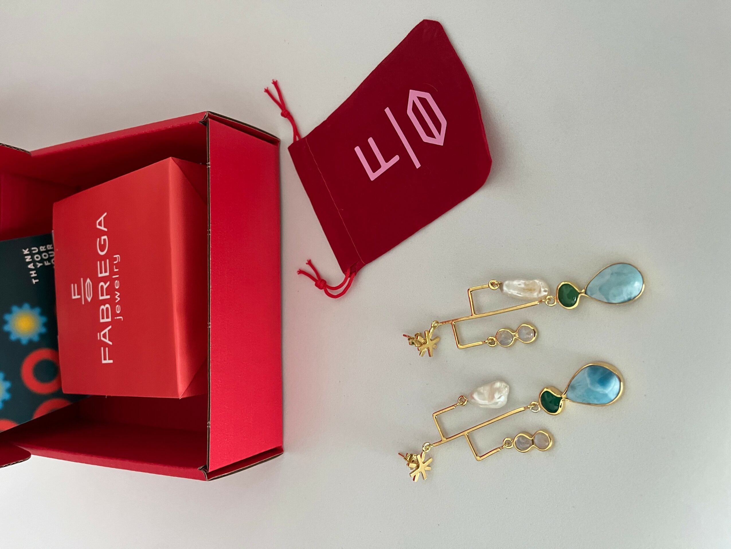 Open red jewelry box with a red pouch and two pairs of earrings with blue and green stones, on a white surface.