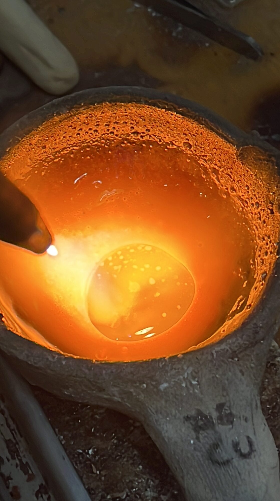 Molten metal glowing orange inside a furnace, with a worker using a tool to handle it.
