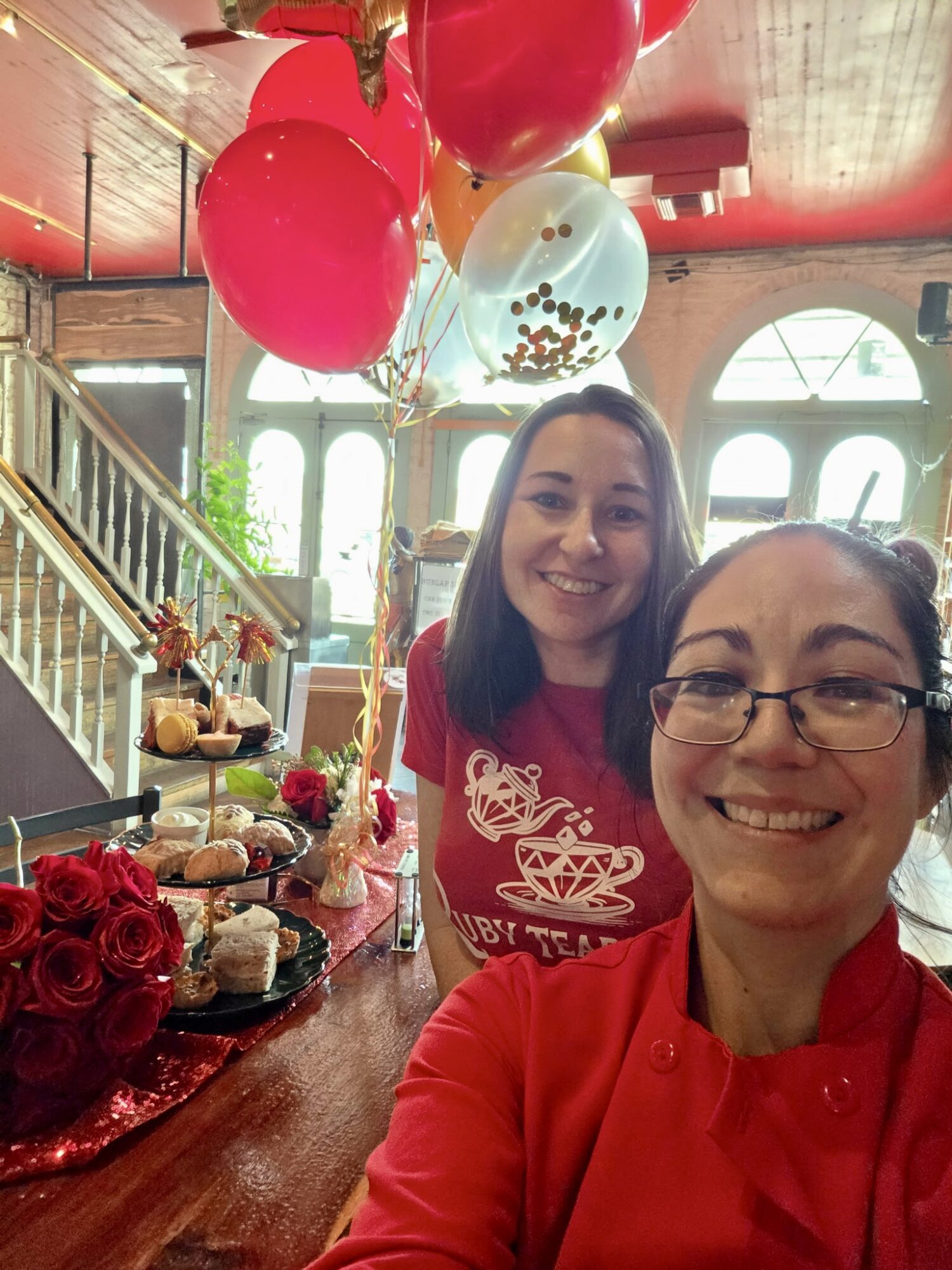 Two women smiling at a celebration with balloons and desserts, one wearing glasses and a red shirt, the other in a red top.