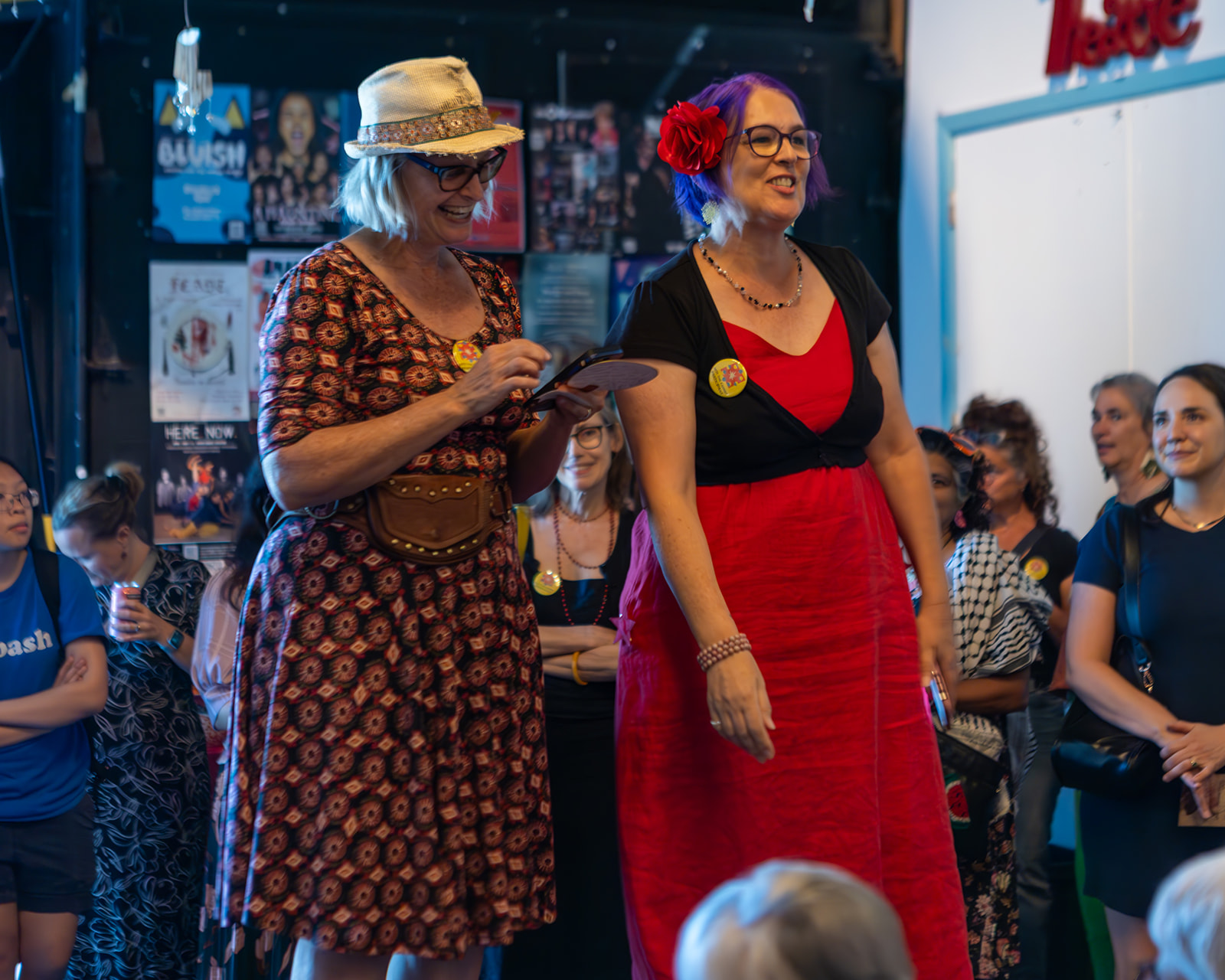 Two women stand together in a crowd, one reading a paper, the other smiling, with posters and people in the background.