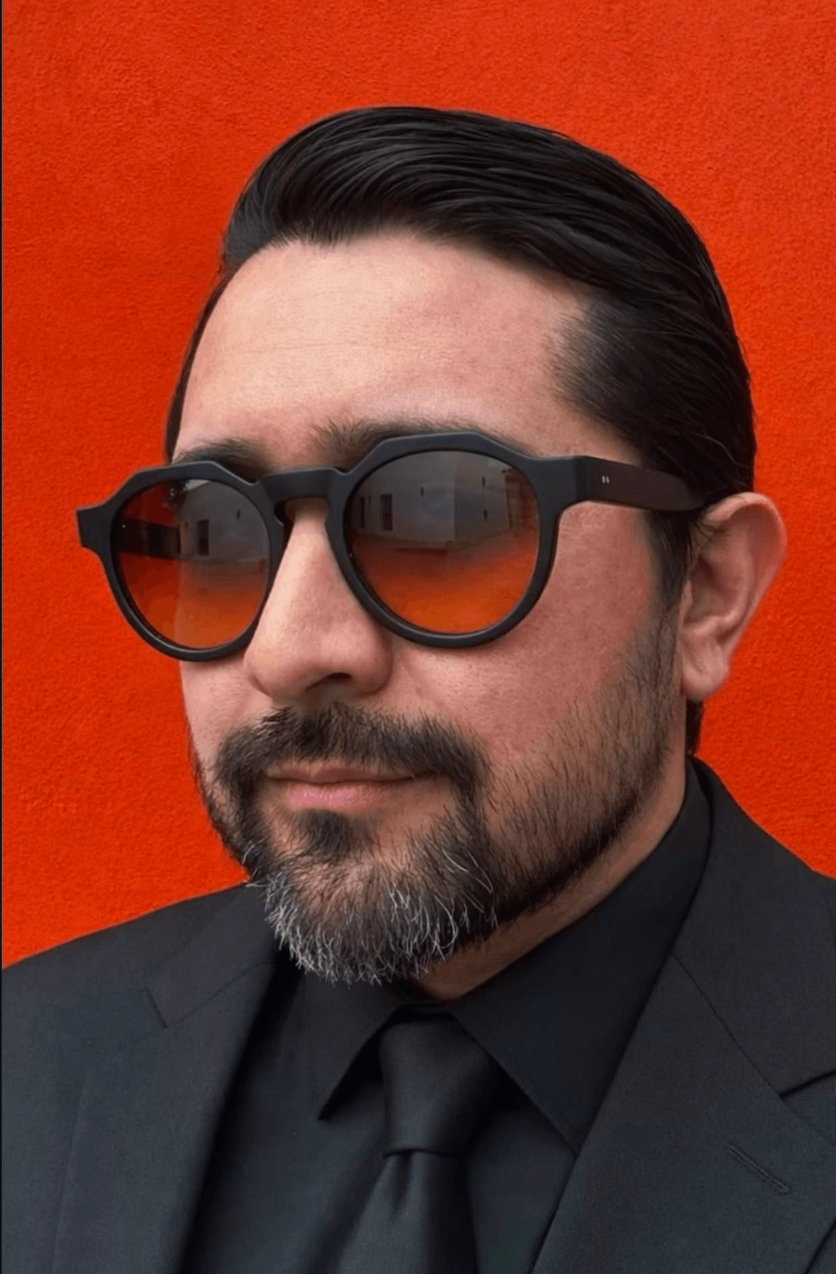 Man with dark hair, beard, sunglasses, wearing a black suit and tie, against an orange background.