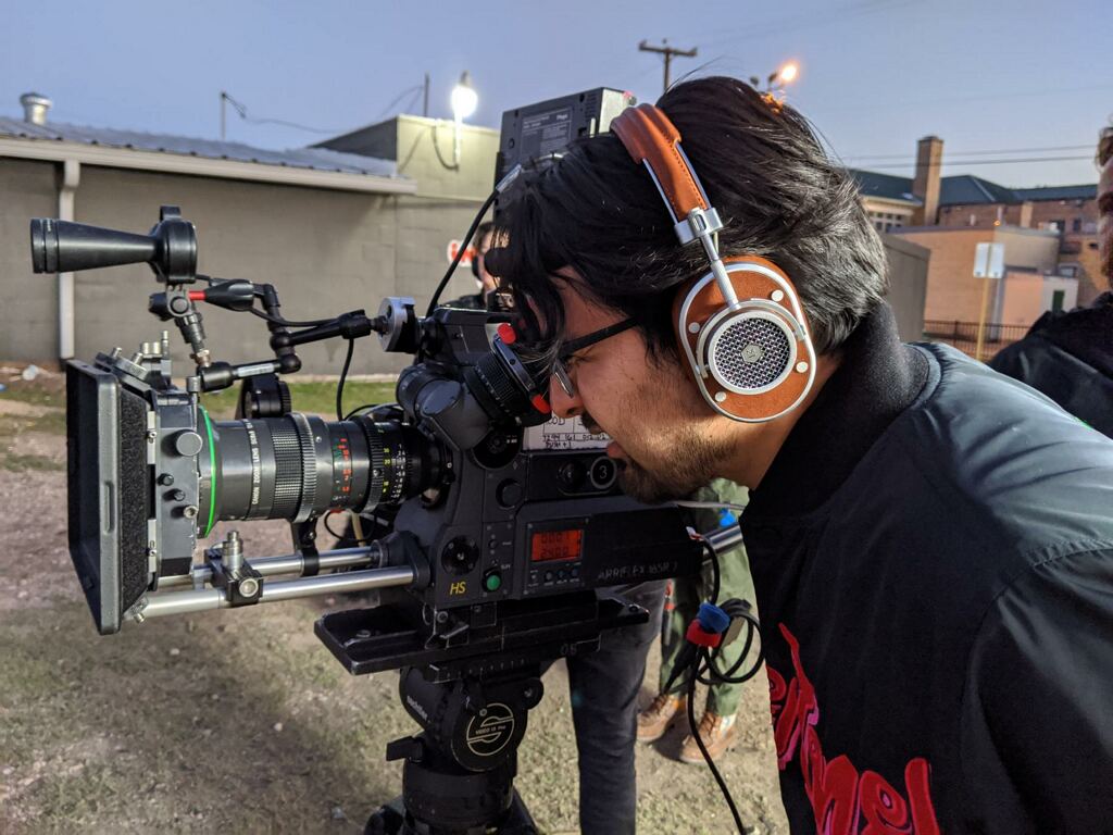Person with headphones operating a professional video camera outdoors, with buildings and power lines in the background.