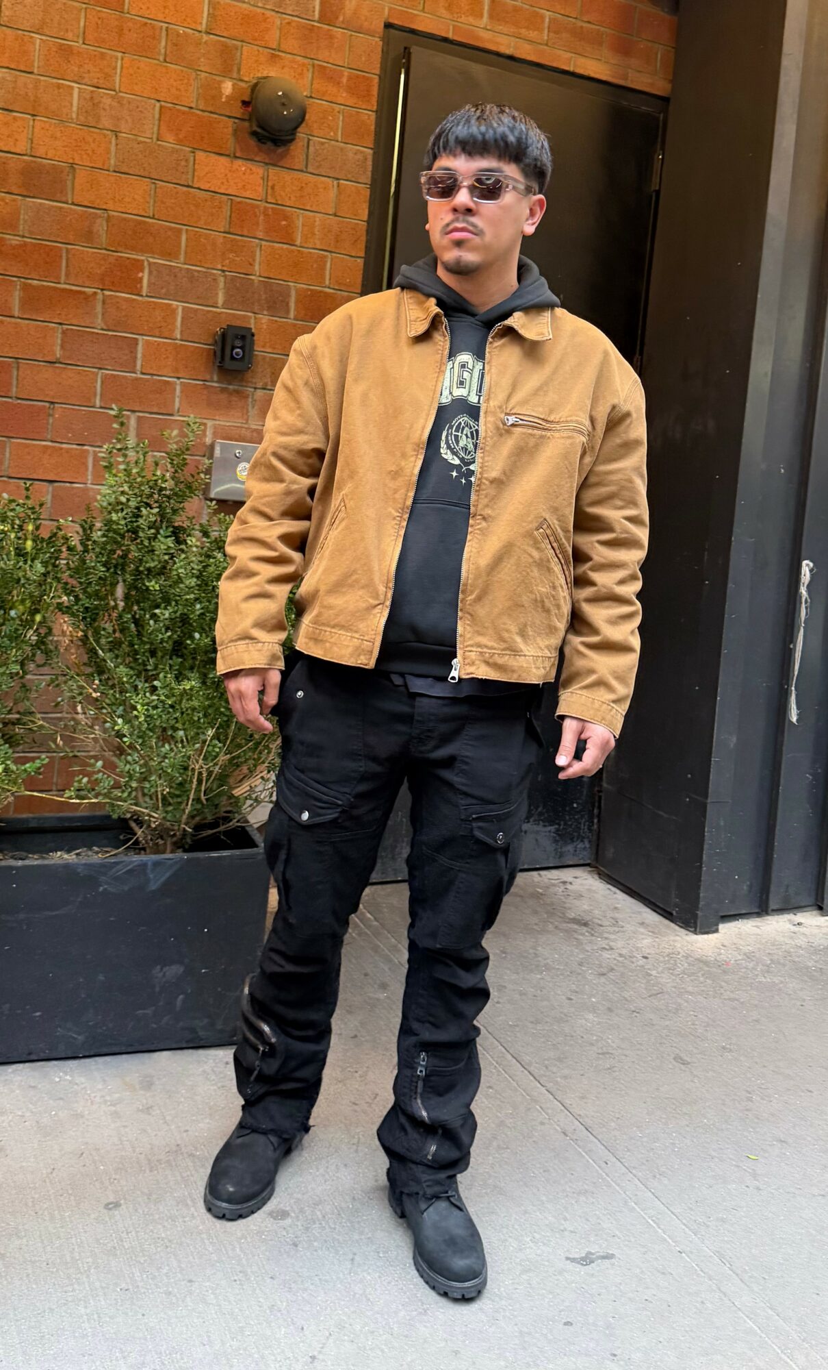 Young man standing outdoors in front of a brick wall and black door, wearing sunglasses, a tan jacket, black hoodie, and black pants.