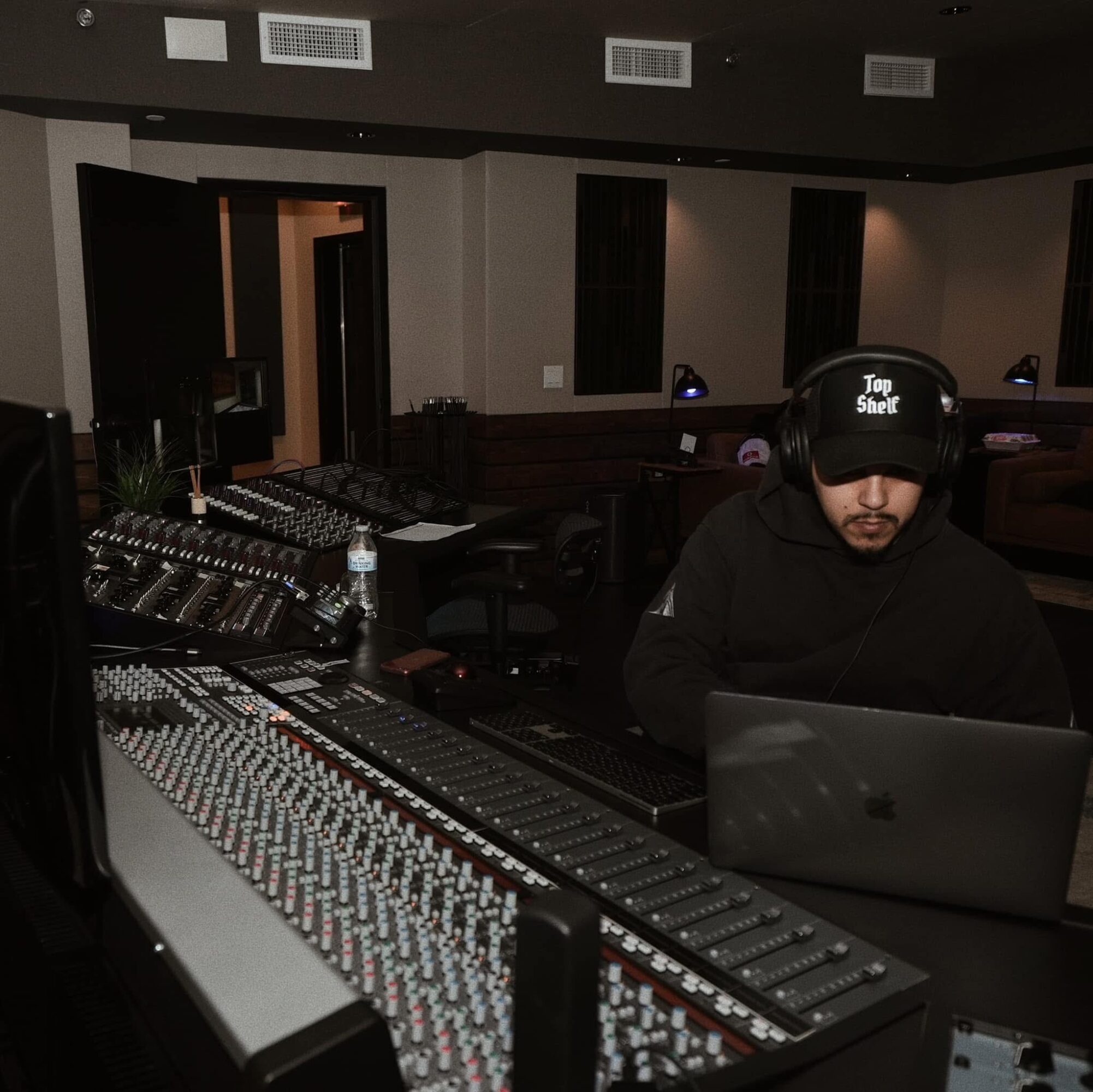 Person wearing a cap with 'Top Shelf' text working on a laptop in a recording studio with audio equipment.