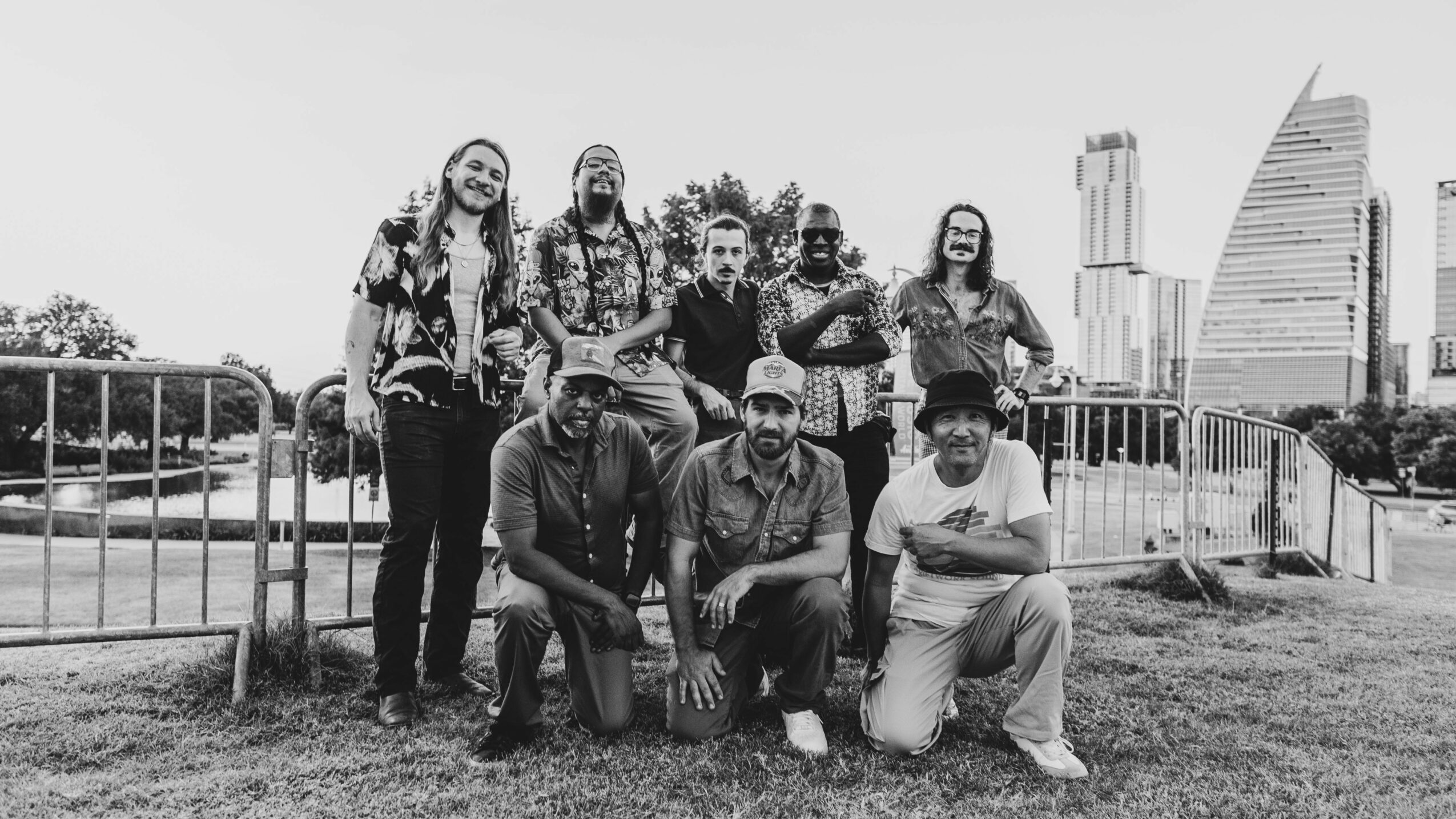 Group of nine people outdoors in front of a city skyline, some standing and some kneeling, with trees and grass.