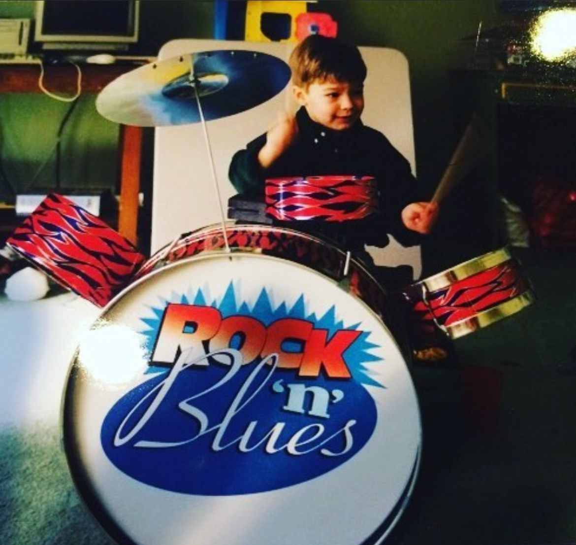Child sitting behind a drum set with a 'Rock 'n' Blues' logo, smiling and holding drumsticks.