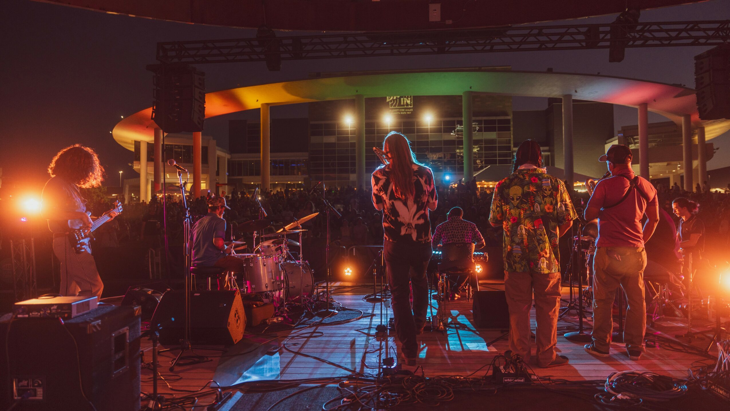 Band performing on stage with colorful lighting at an outdoor event, audience in front, evening setting.
