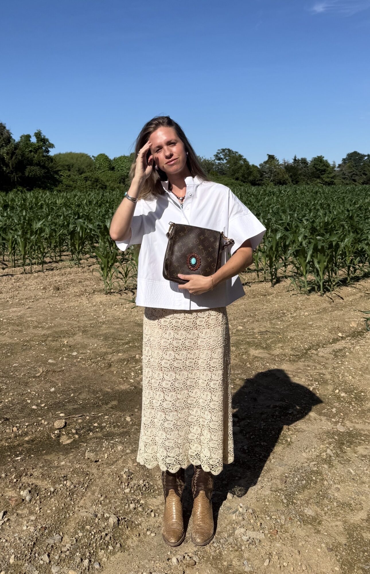 Woman standing outdoors in a field with green plants, wearing a white top and beige skirt, holding a bag, under a blue sky.