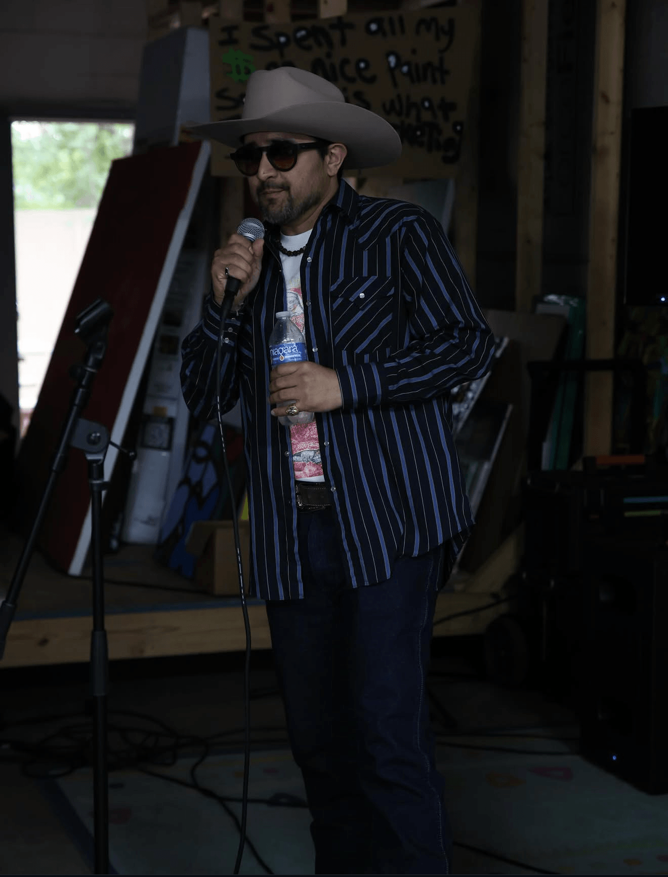 Person wearing a large hat, sunglasses, striped jacket, holding a microphone and water bottle, standing indoors with equipment.