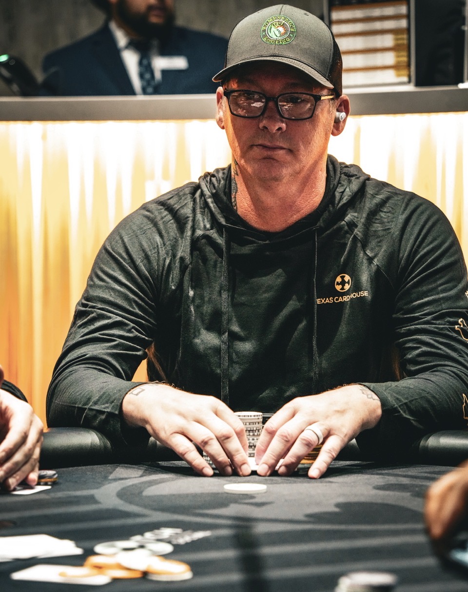 Person wearing glasses and a cap, sitting at a poker table with chips, in a casino setting.