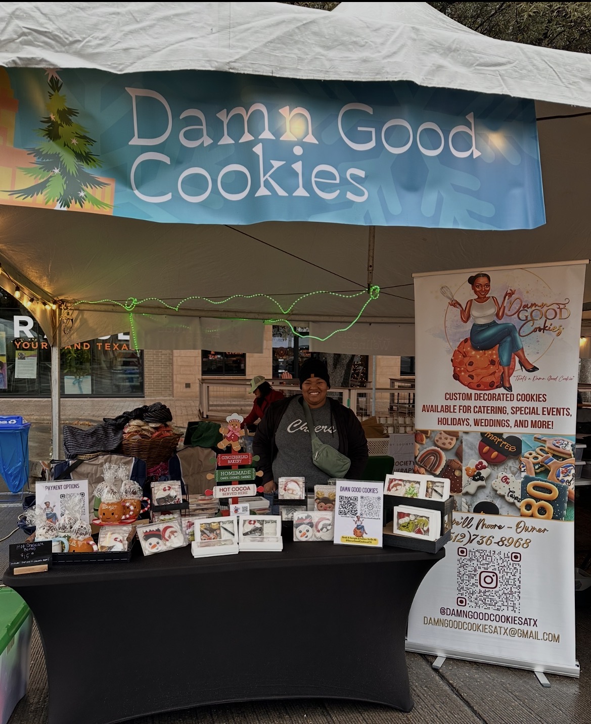 Vendor stand with cookies and a sign that reads 'Damn Good Cookies' under a canopy, with a person behind the table.