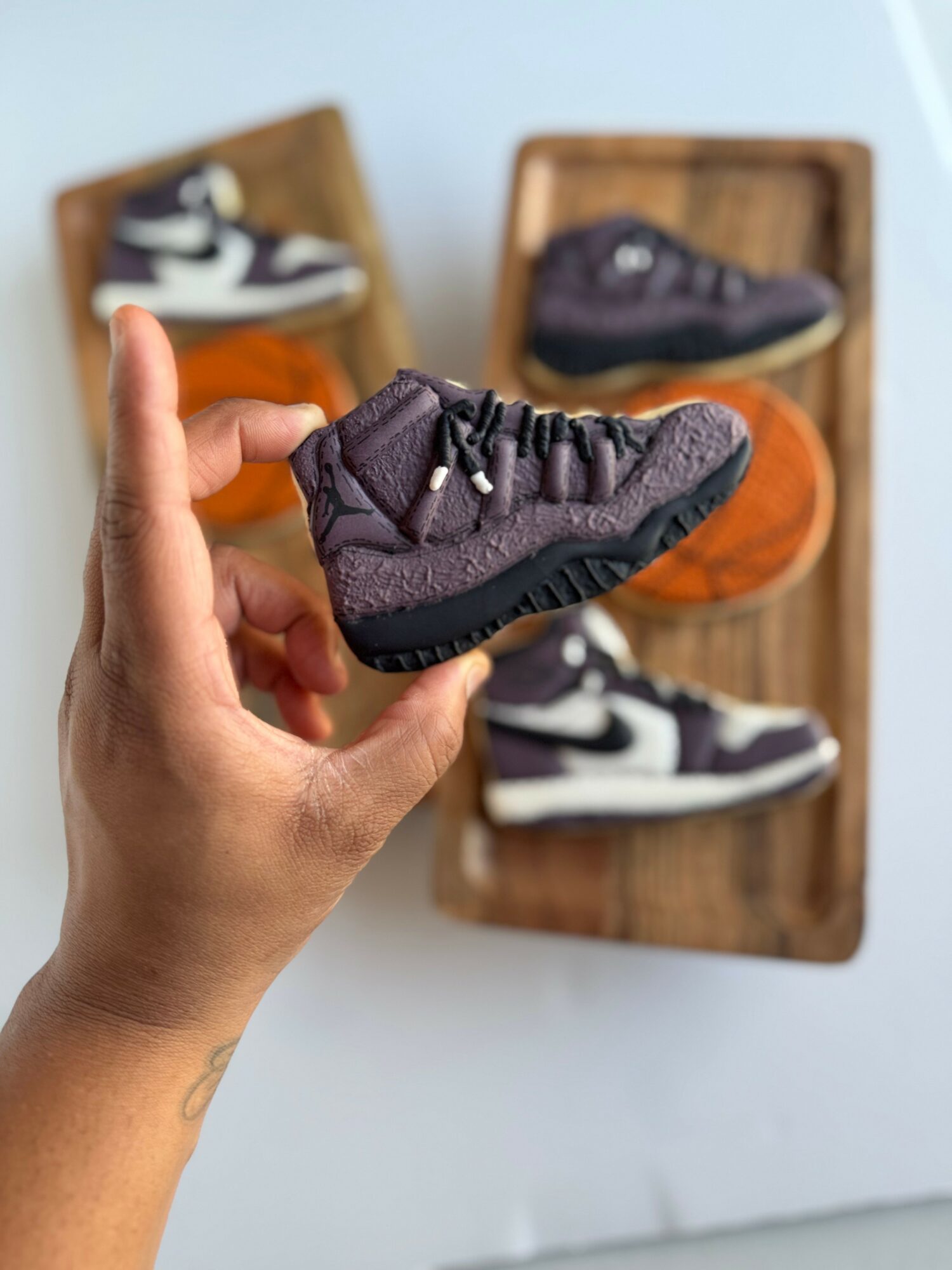Hand holding a small black sneaker with a textured sole, with two other sneakers displayed on wooden shelves in the background.