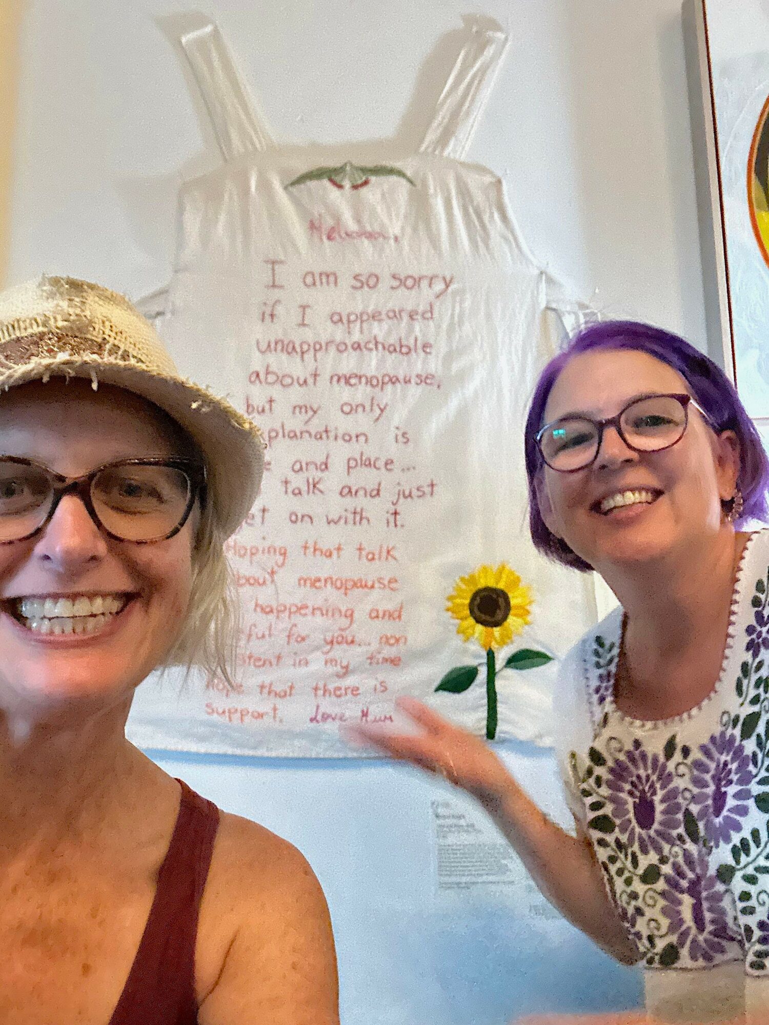 Two women smiling in front of a wall with a handwritten note and a sunflower drawing, one wearing glasses and a hat.