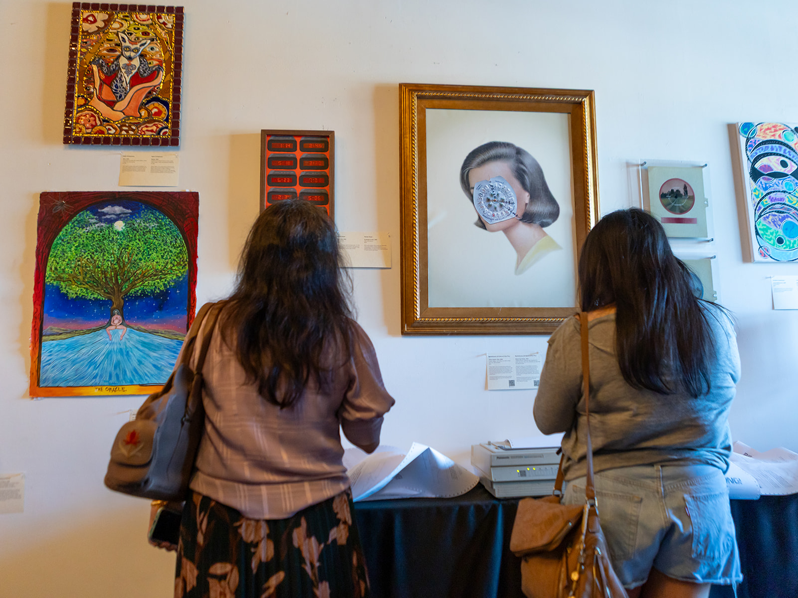 Two women with long dark hair look at artwork on a gallery wall, including a framed portrait and colorful paintings.