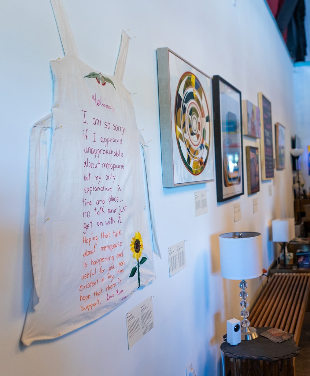 Canvas bag with handwritten message and sunflower, displayed on a wall with framed artwork and a lamp below.