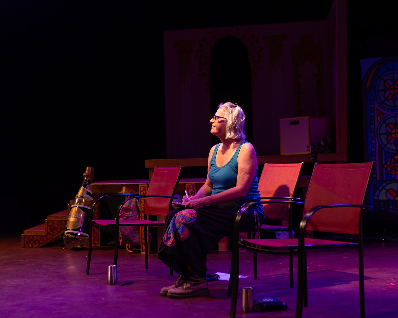 Woman with gray hair sitting on a stage, surrounded by chairs and props, with a dark background and colorful lighting.