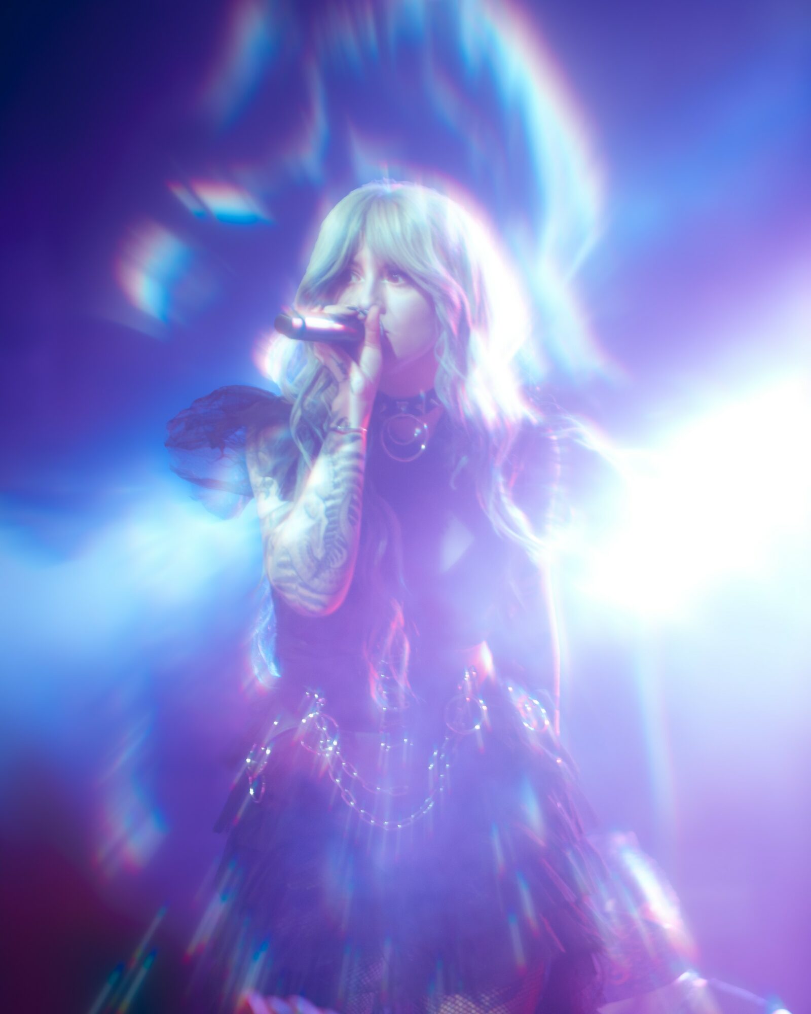 Performer with light-colored hair singing into a microphone on stage with colorful, blurred lighting effects