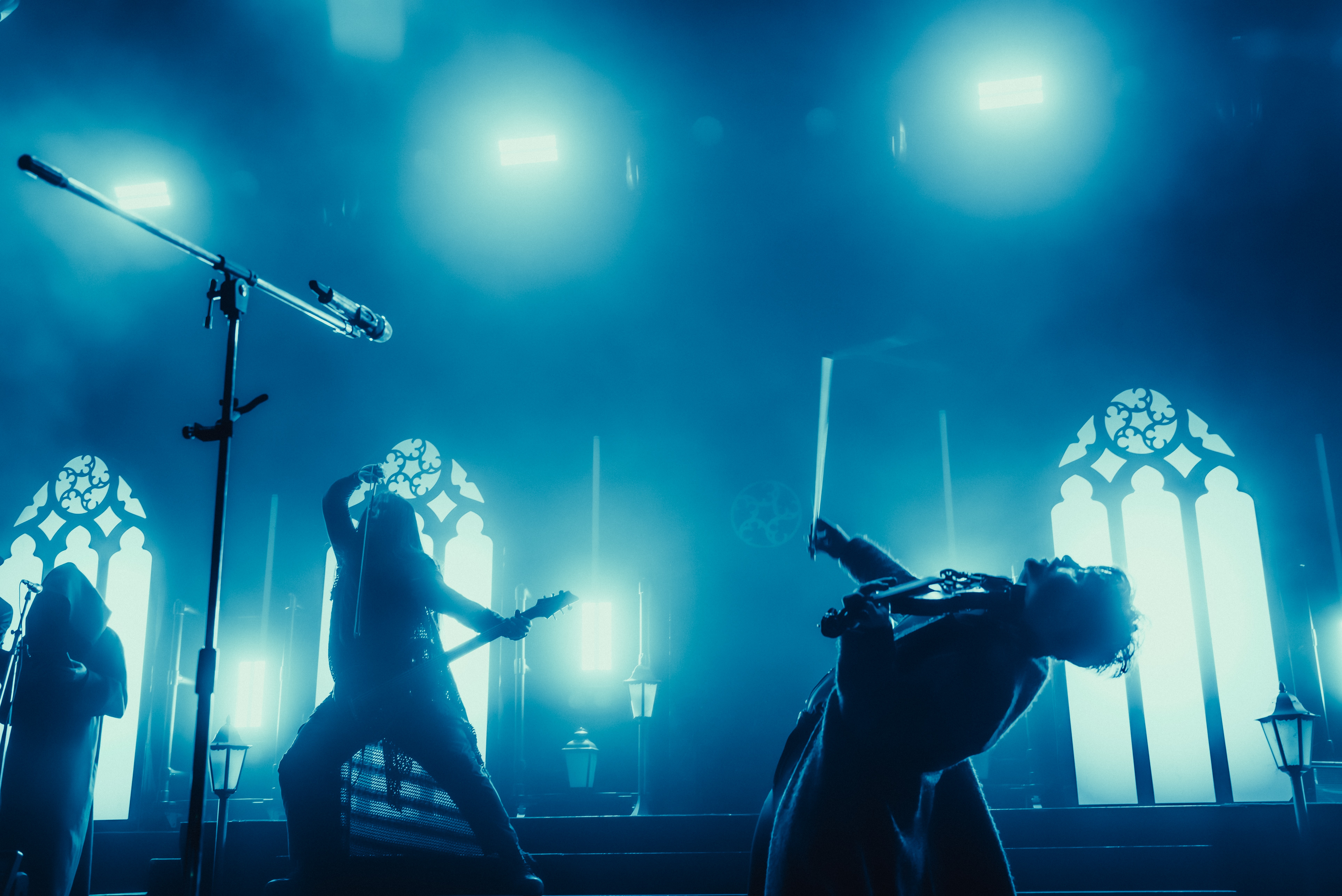 Band performs on stage with bright blue lighting and gothic-style windows in the background.