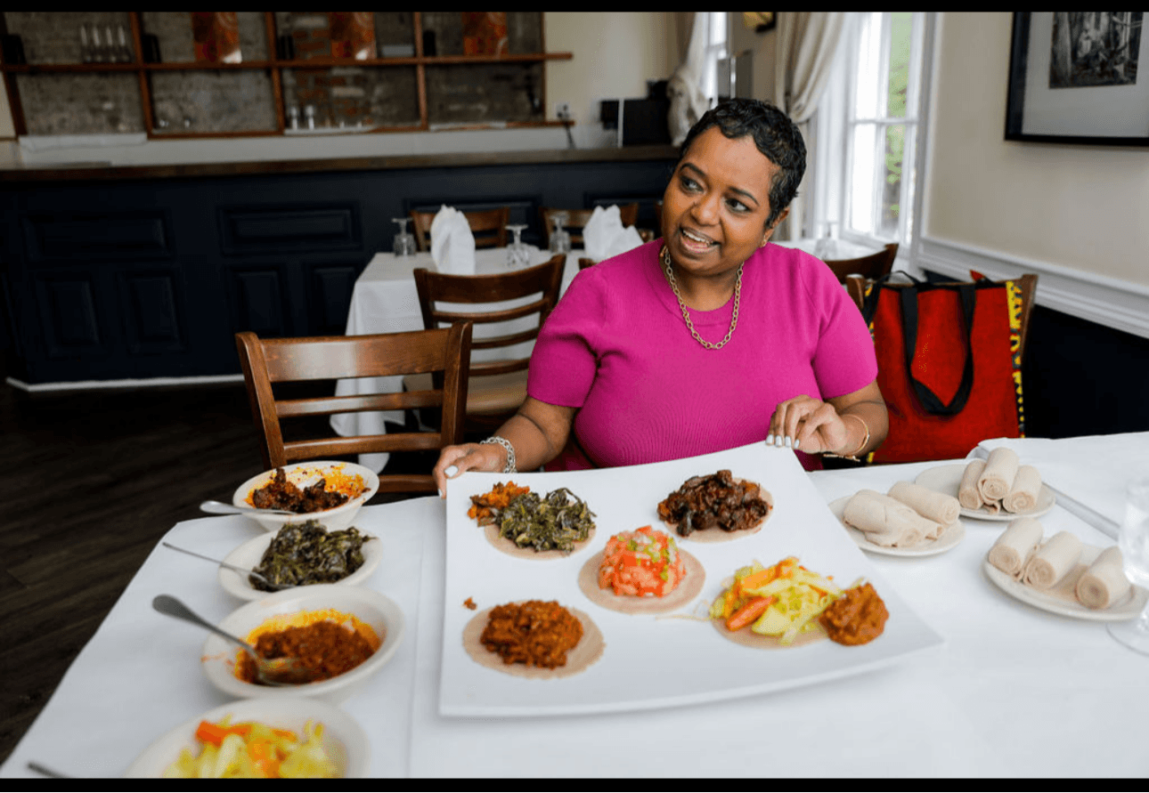 Woman in pink shirt sitting at a table with a large platter of various foods, smiling and looking to her right.