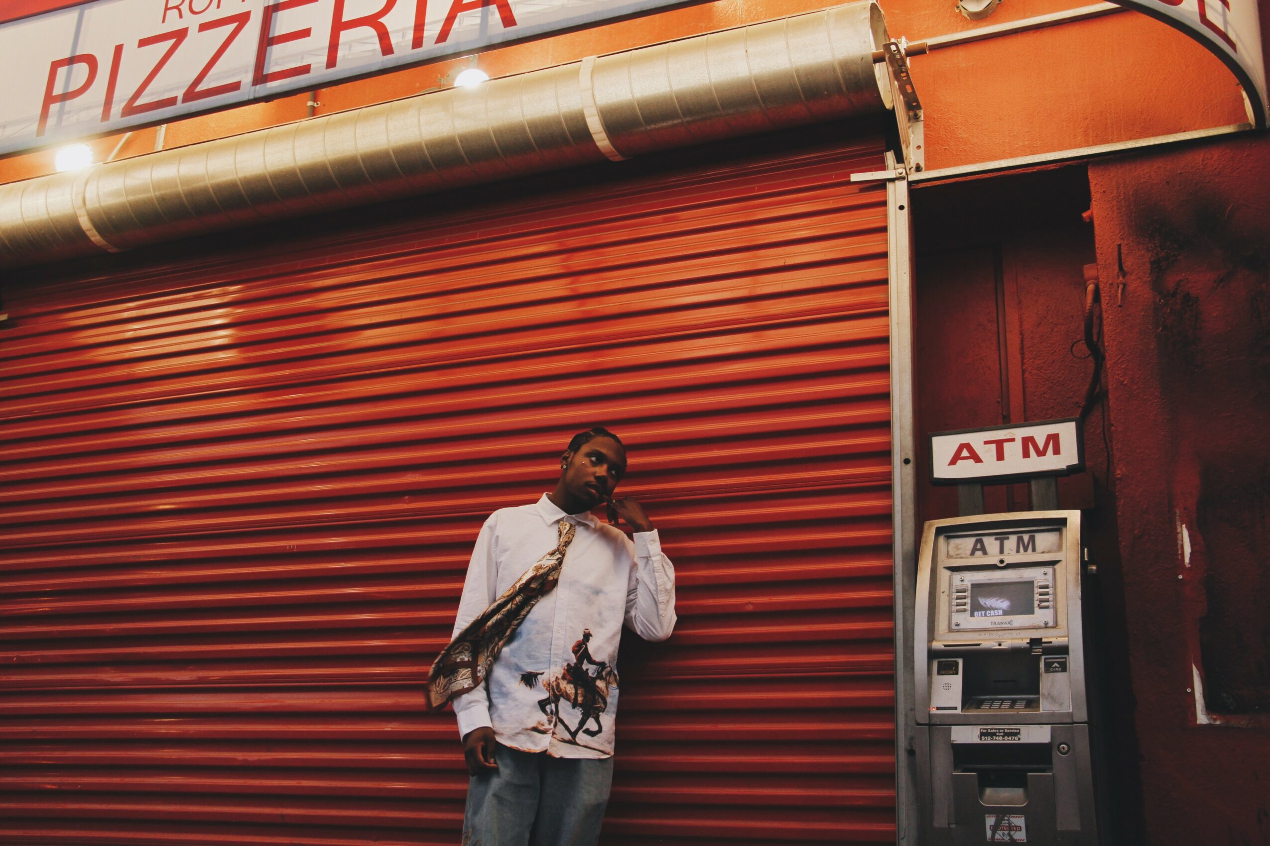Person standing in front of a red closed shutter with an ATM nearby, holding a bag and wearing sunglasses.