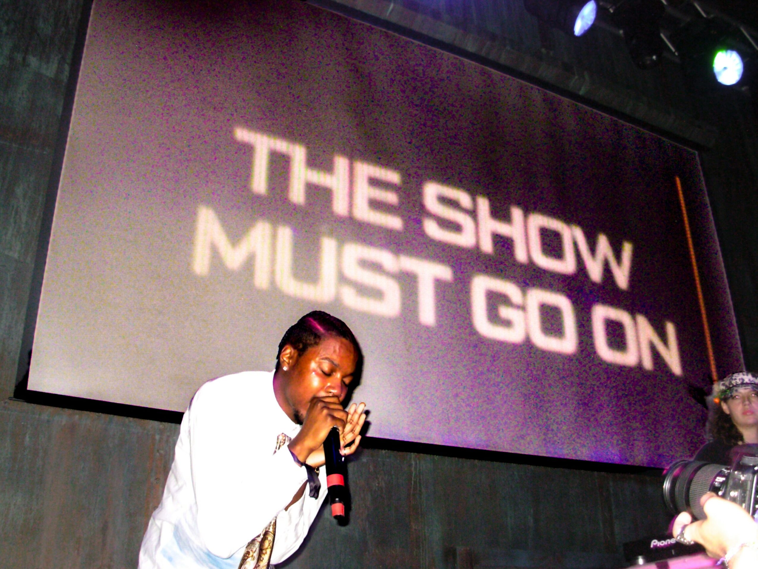 Person singing into a microphone on stage with a large screen behind displaying text.