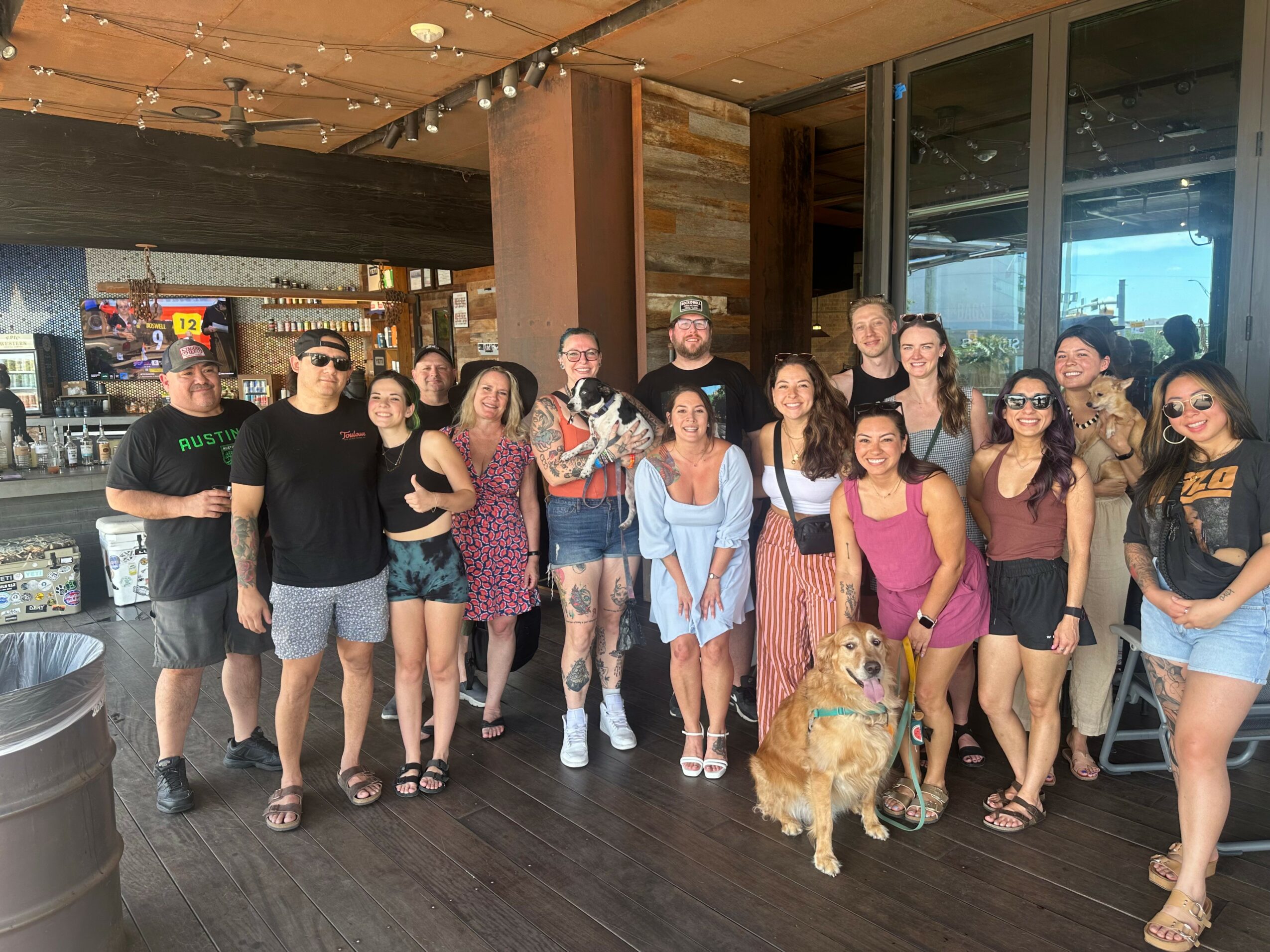 Group of people standing indoors near large windows, with a dog in front, some wearing sunglasses and casual clothing.