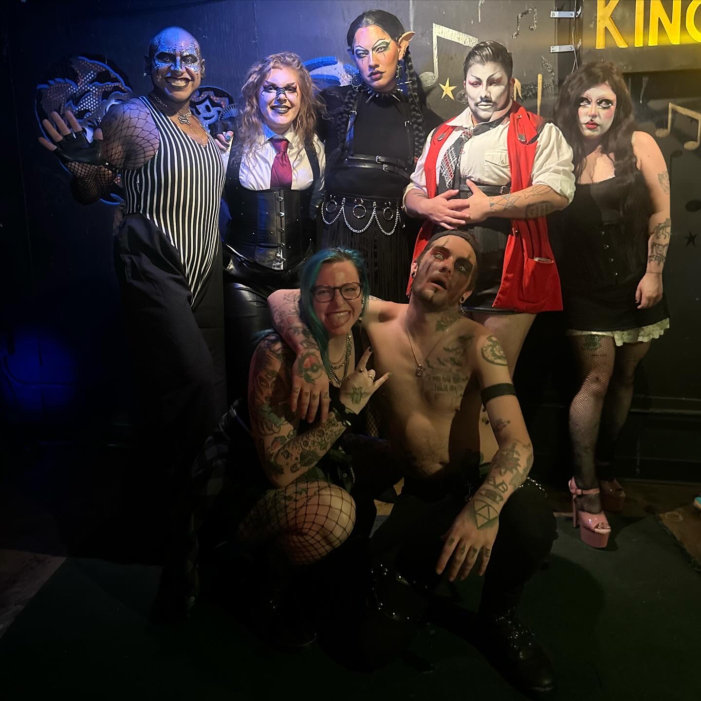 Group of seven people, some with makeup and costumes, posing together on stage with dark background and stage lights.