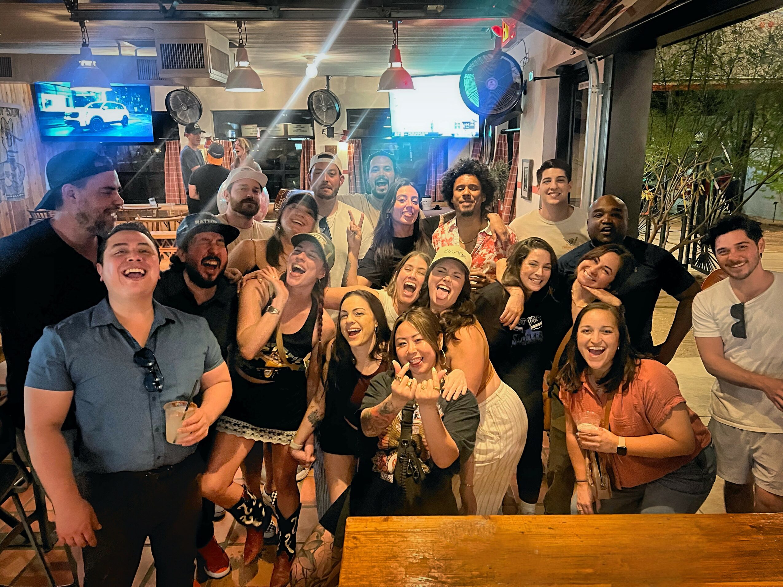 Group of people smiling and celebrating indoors with drinks, some raising hands, in a lively social setting.