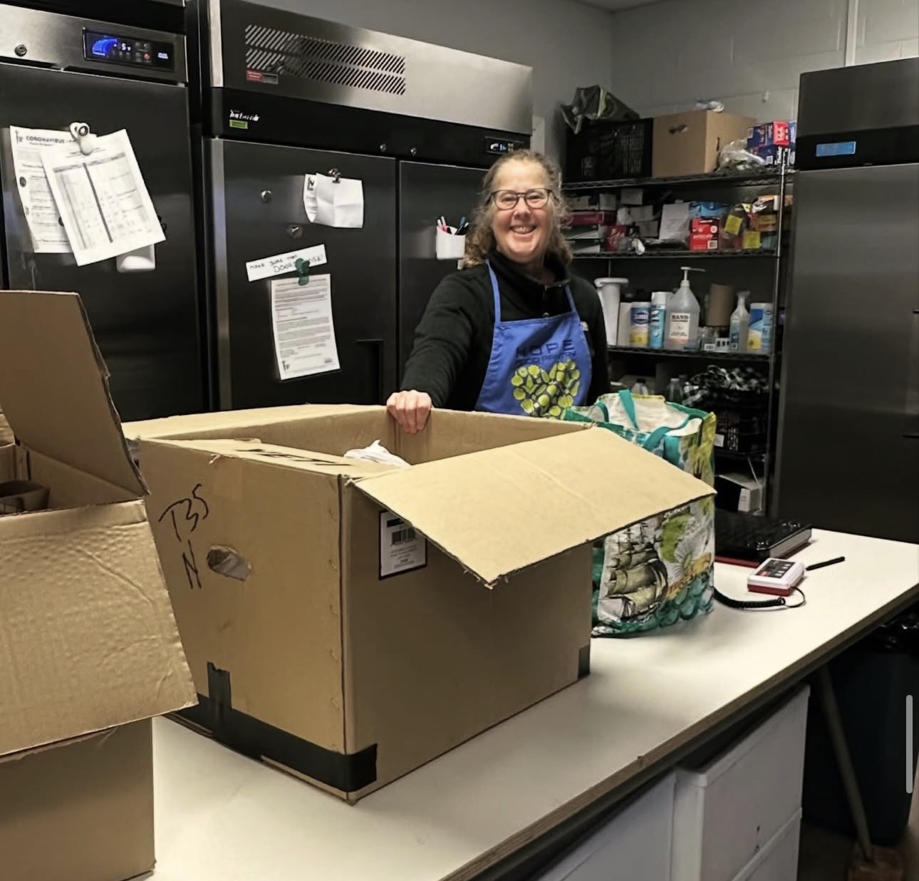 Smiling woman in a blue apron stands behind a table with a large open cardboard box, in a storage room.