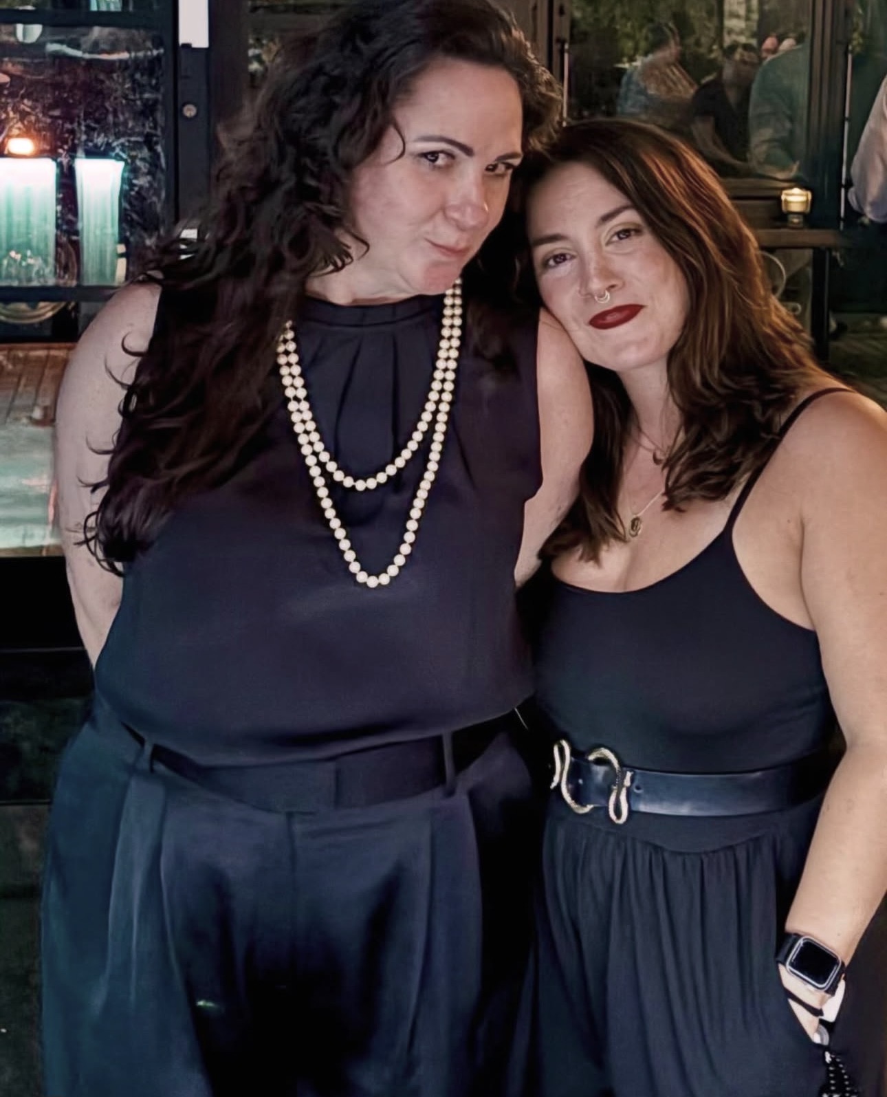 Two women standing close together, one with dark curly hair and pearl necklace, the other with straight hair and red lipstick, in a dimly lit setting.