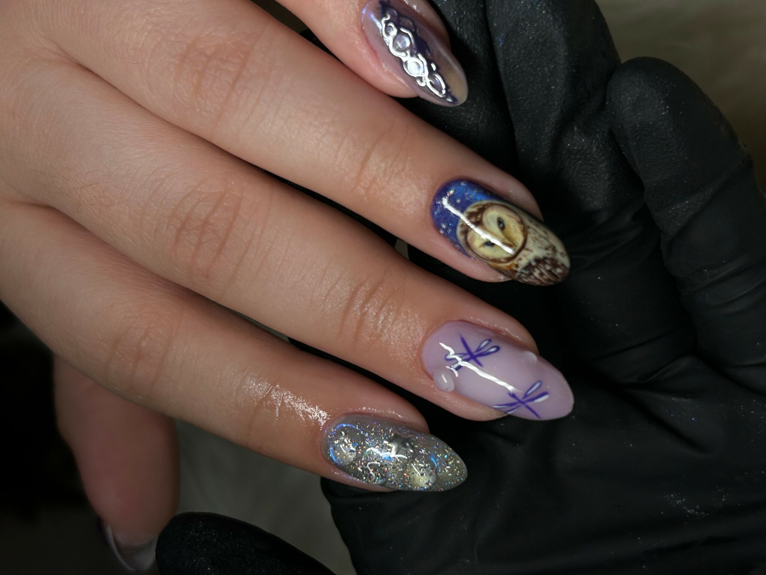 Close-up of a hand with decorated nails, including silver glitter, purple, and gold designs, held by a gloved hand.
