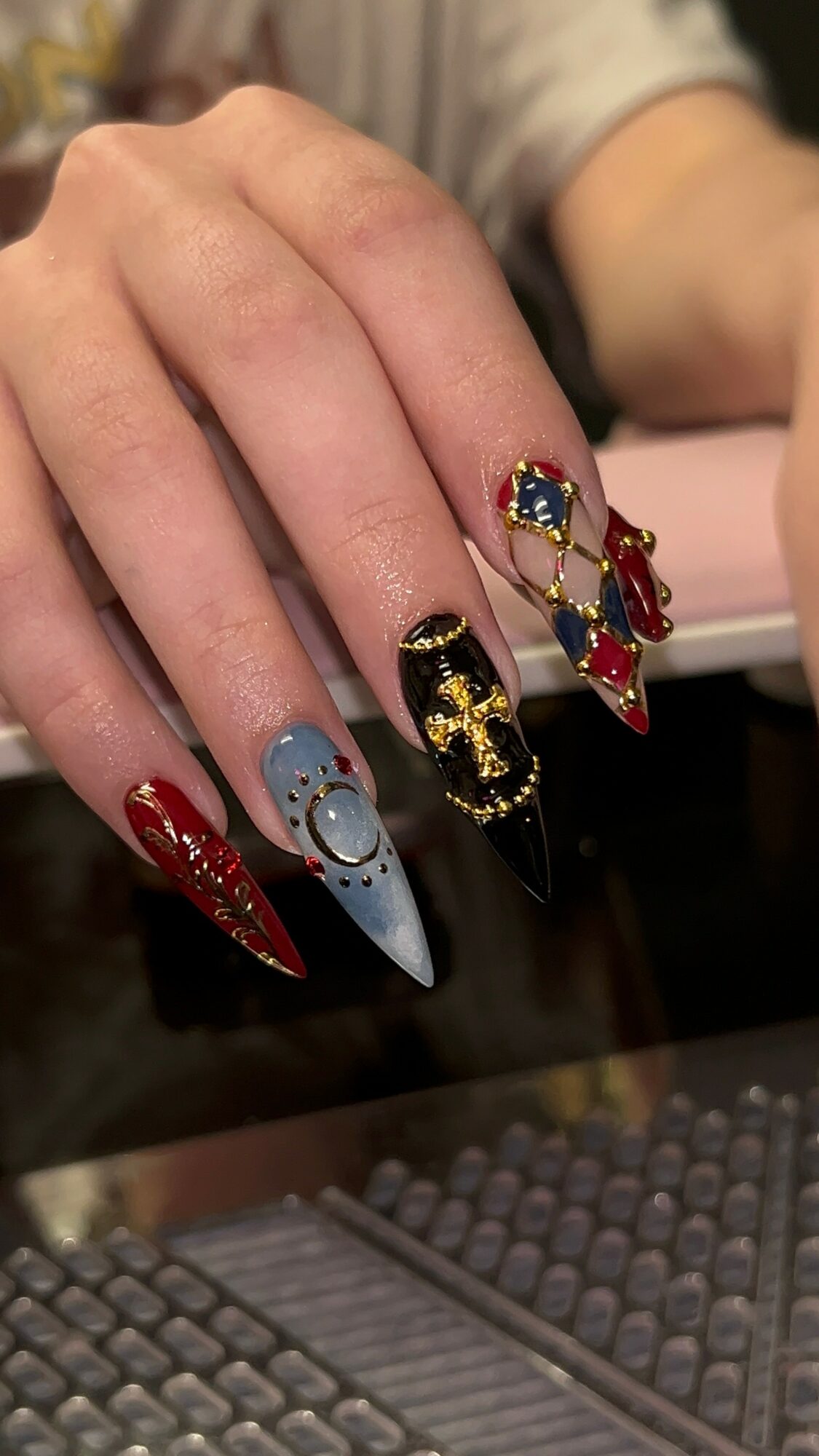 Hand with decorated nails featuring red, black, and blue colors, gold accents, and rhinestones, resting on a surface.