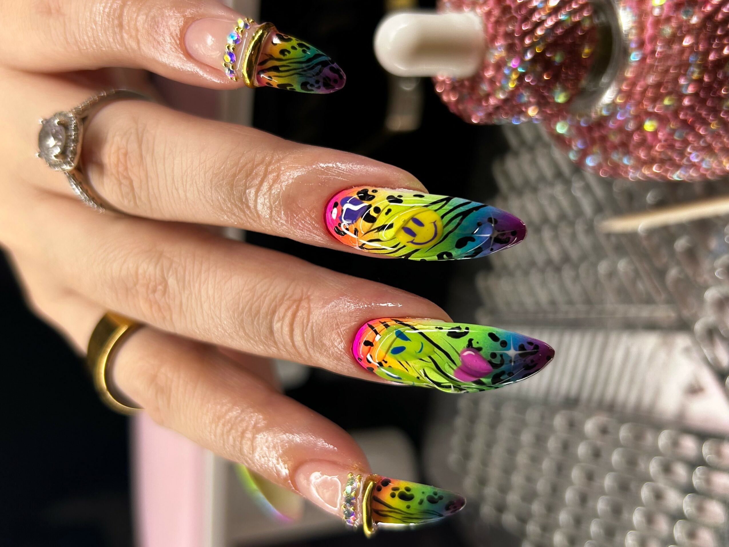 Hand with long, colorful, floral and abstract nail art, rings on fingers, background with pink and glittery objects.
