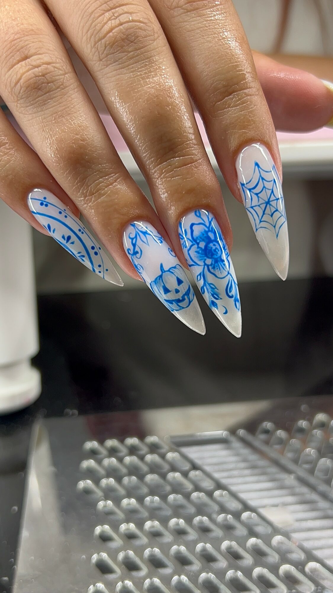 Hand with decorated nails featuring blue and white snowflake and web designs, long pointed shape, skin tone visible.