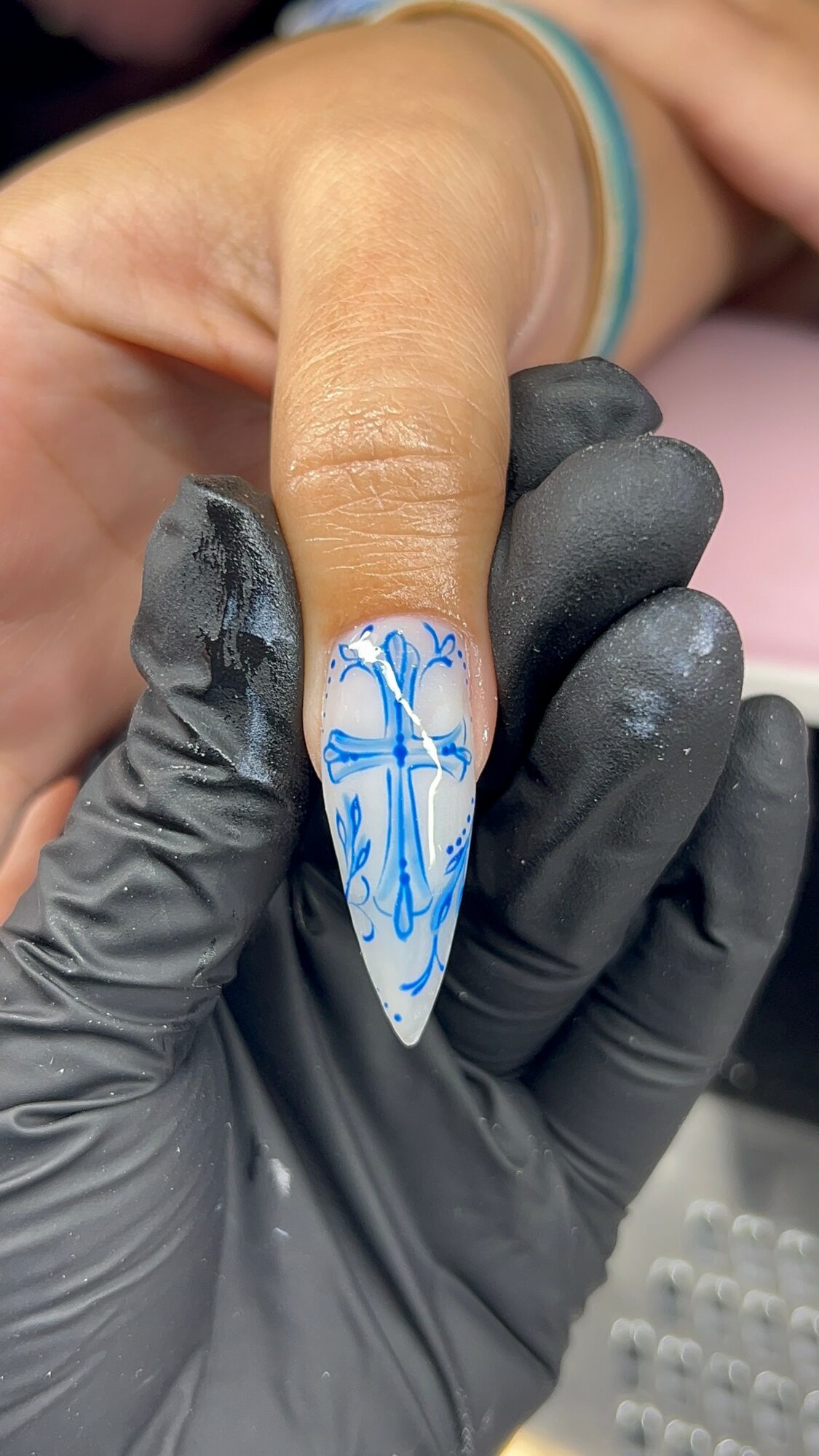 Close-up of a hand with a decorated fingernail featuring blue and white nail art, held by a person wearing black gloves.