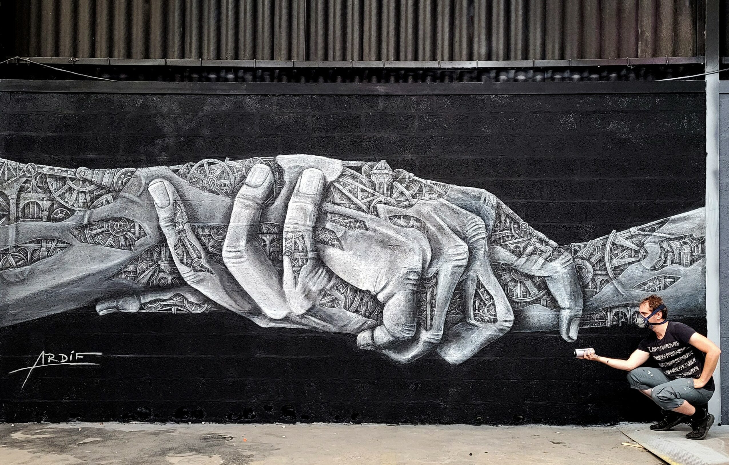 A person crouches next to a large black and white mural of two hands clasped together, painted on a wall.