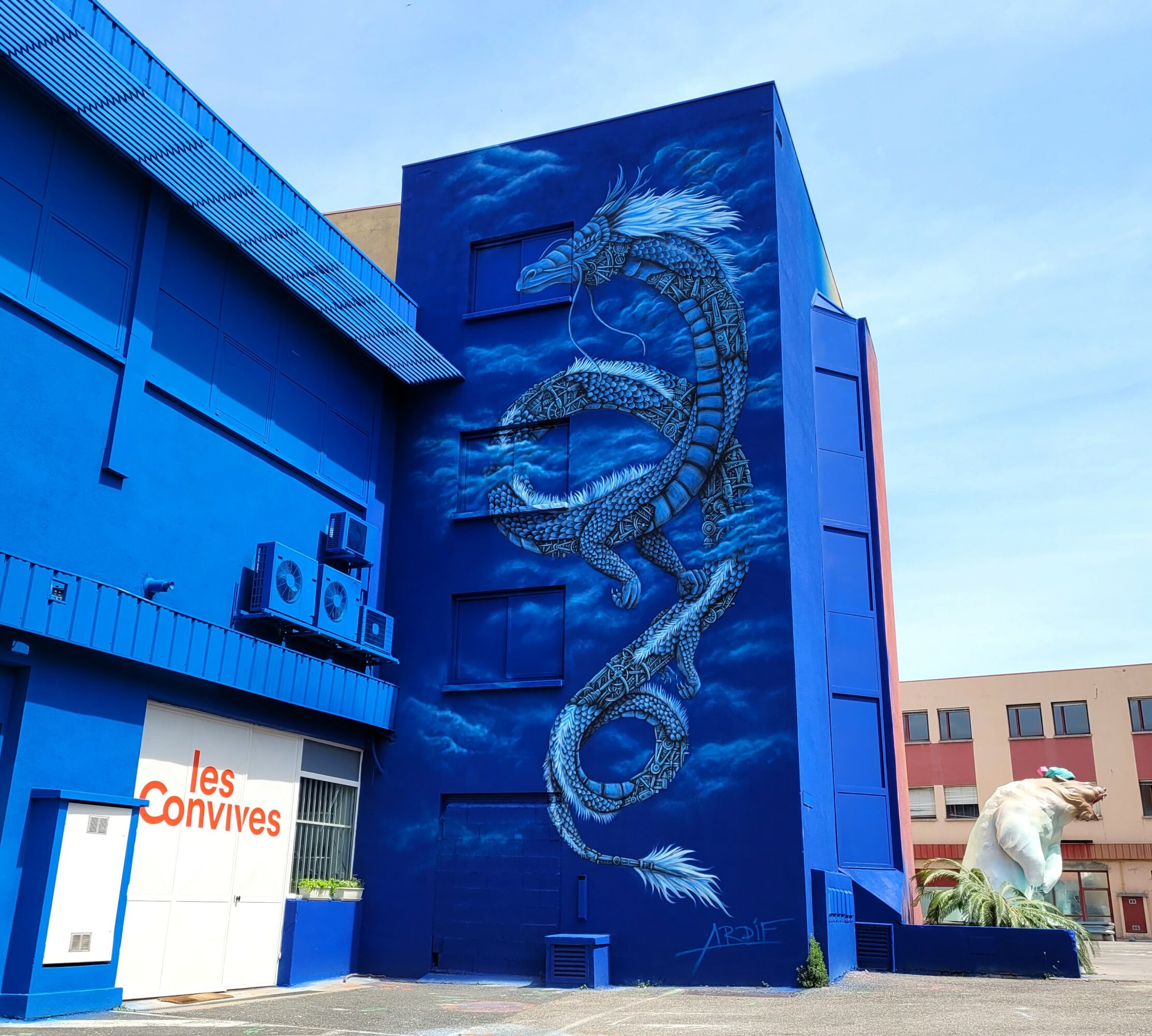 Blue building with a large dragon mural, and a sign reading 'les convives' on the lower left.