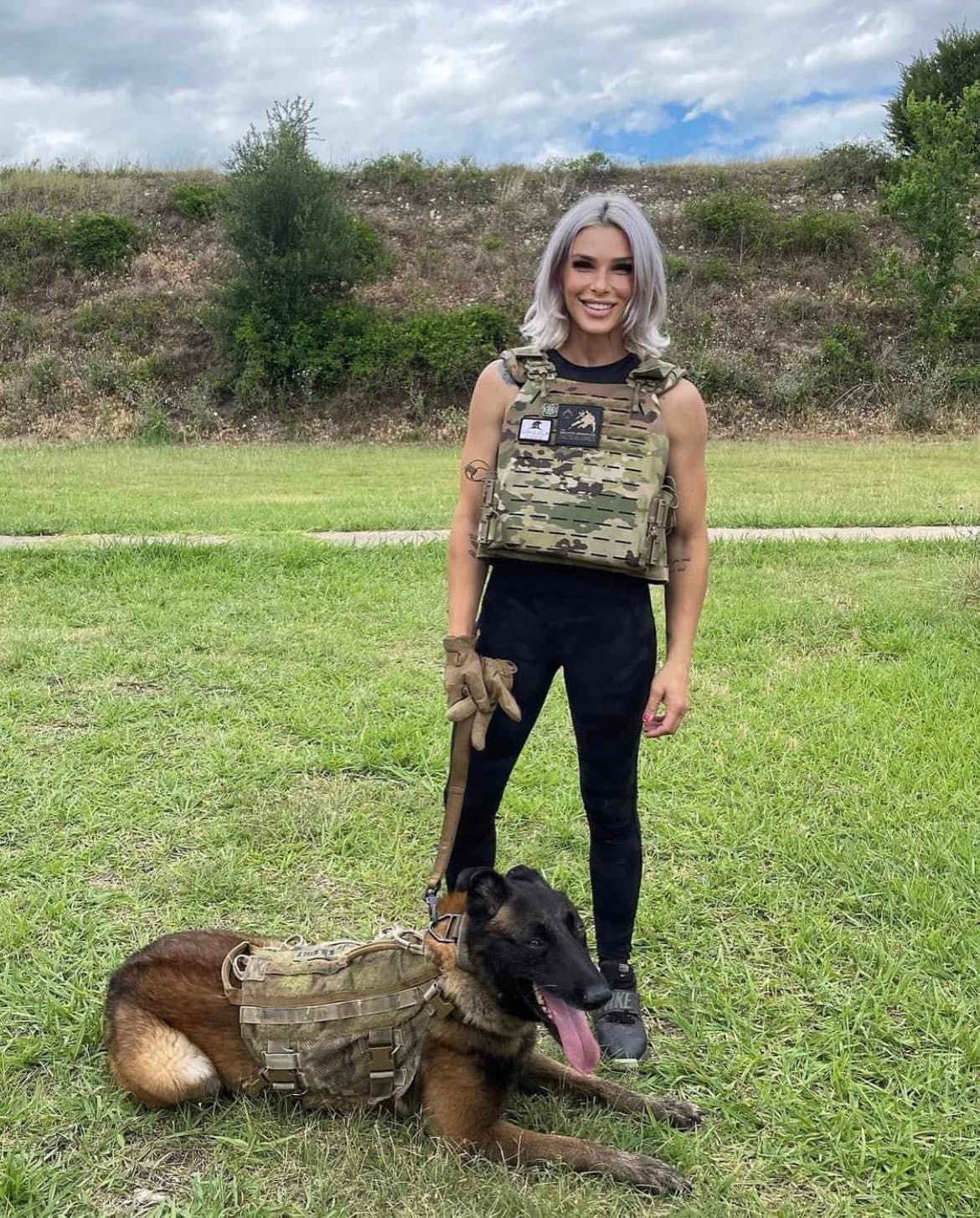 Woman in military vest standing on grass with a dog lying down, both wearing tactical gear, outdoors with trees and sky.