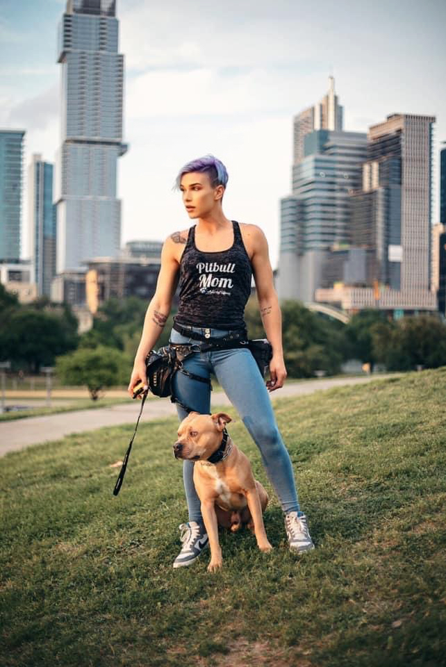 Person with short purple hair standing on grass with a dog, city skyline in background, daytime.