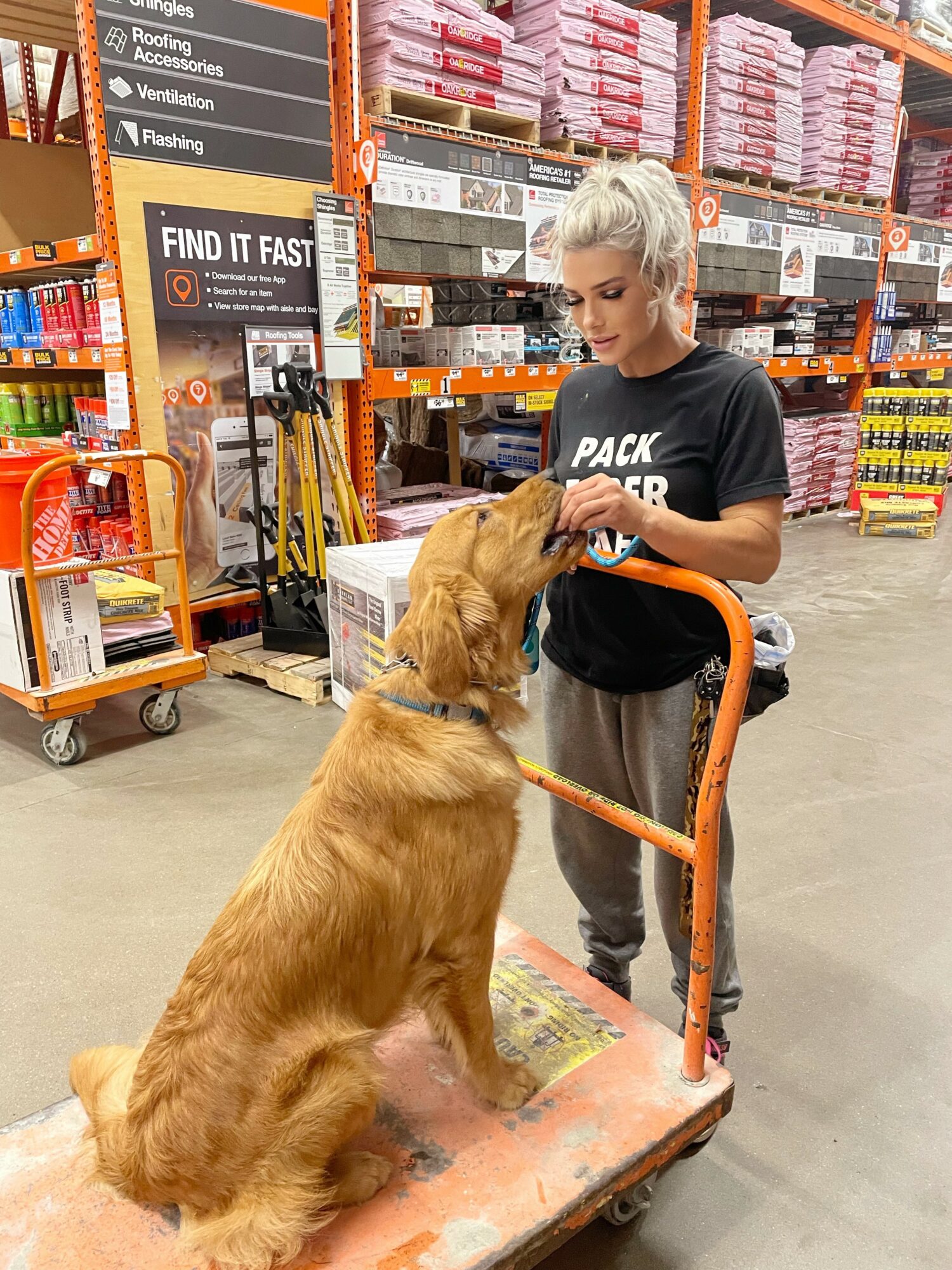 Young woman with platinum hair and a black shirt interacts with a golden retriever in a store aisle, pushing a shopping cart.