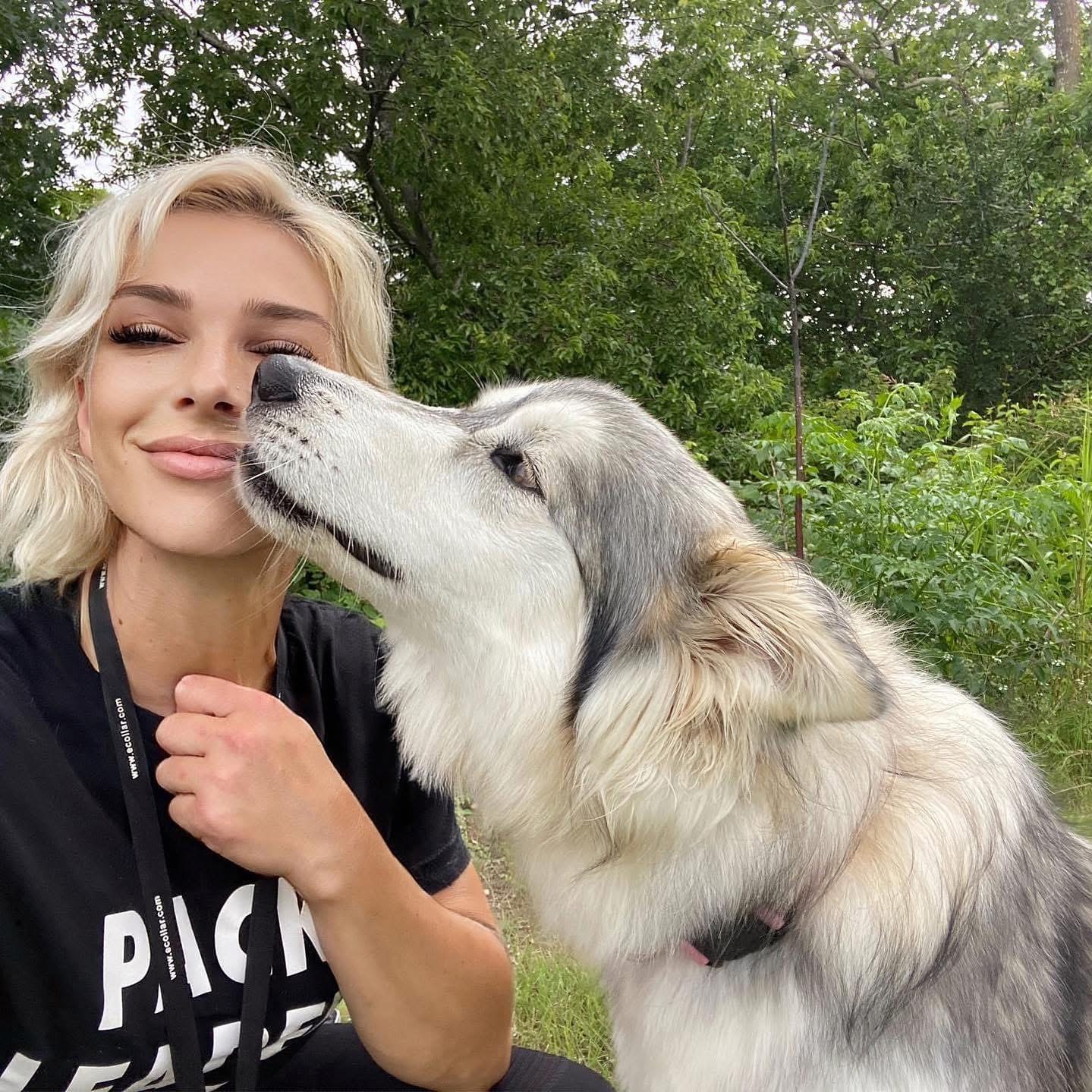 Young woman with blonde hair smiling as a large dog licks her face outdoors with green trees in background.