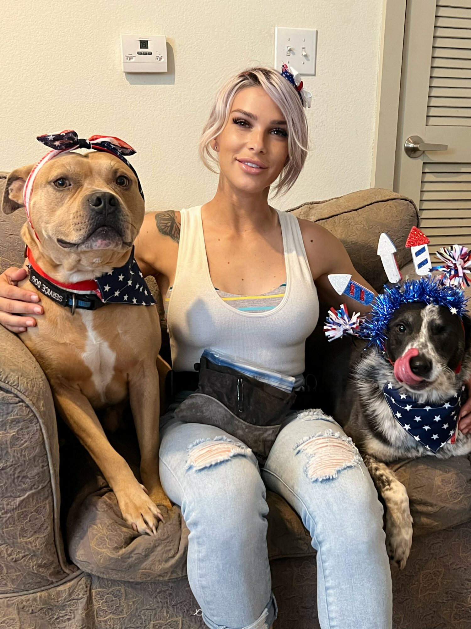 Woman sitting on a couch with two dogs, one wearing a bow and the other with holiday headgear, in a living room.