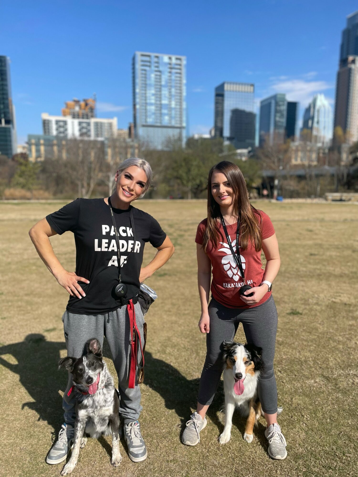 Two women stand outdoors with dogs, city skyline in background, sunny day, both smiling, casual clothing, grassy area.