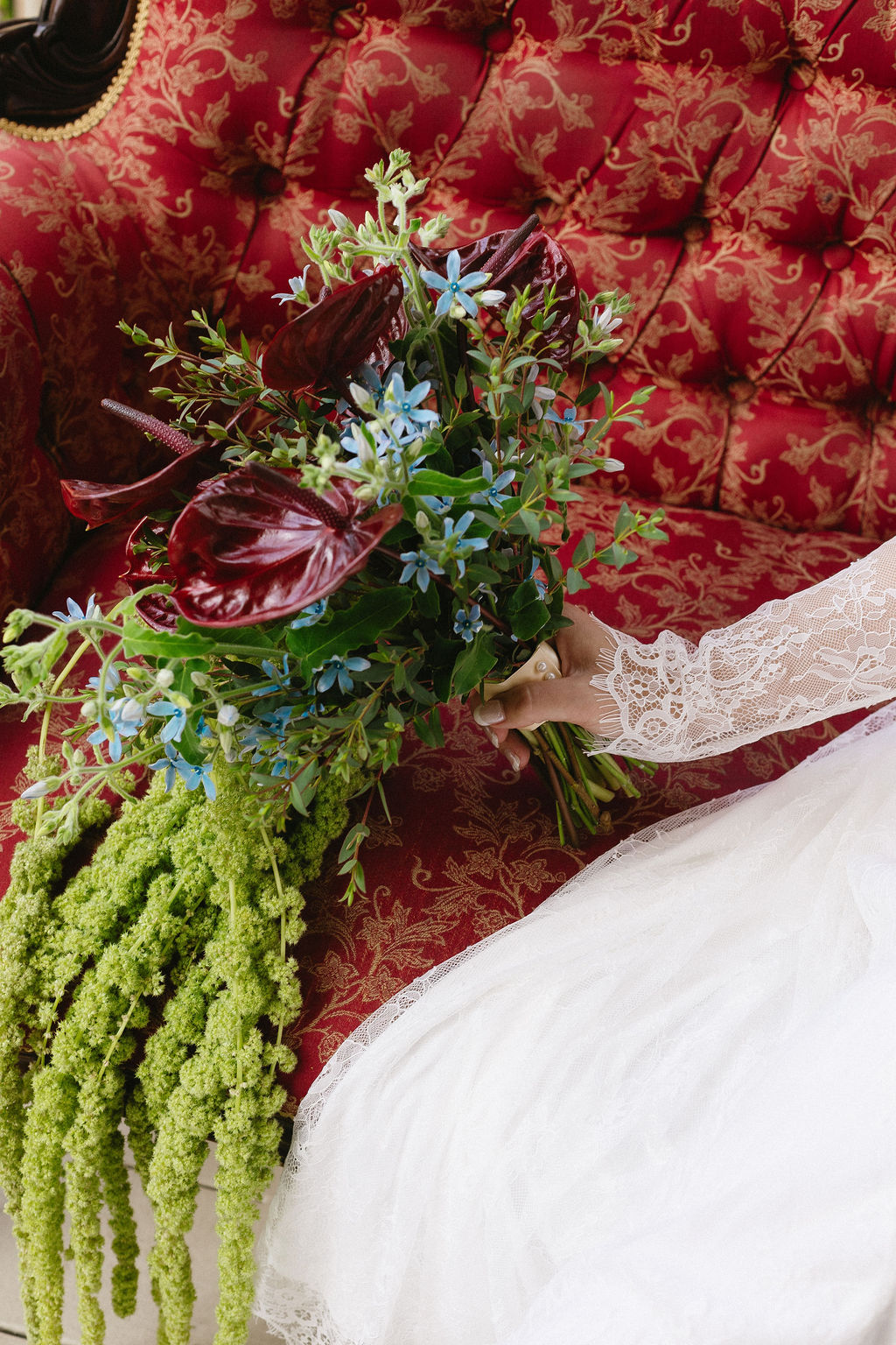 Person in white dress holding a bouquet of dark red, blue, and green flowers and greenery, sitting on a red patterned sofa.