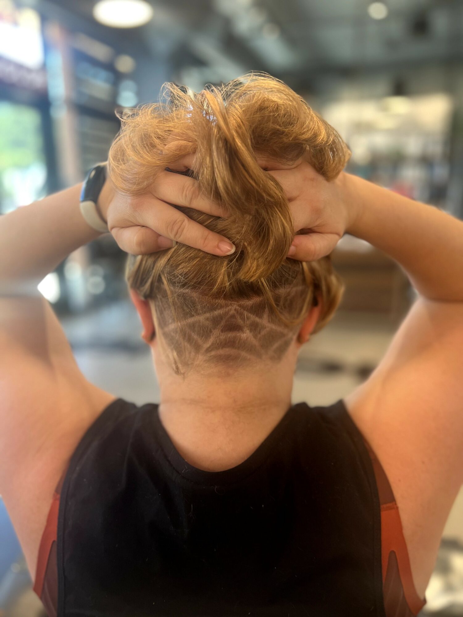 Person with short hair and a shaved design on the back of their head, holding their hair up with both hands.