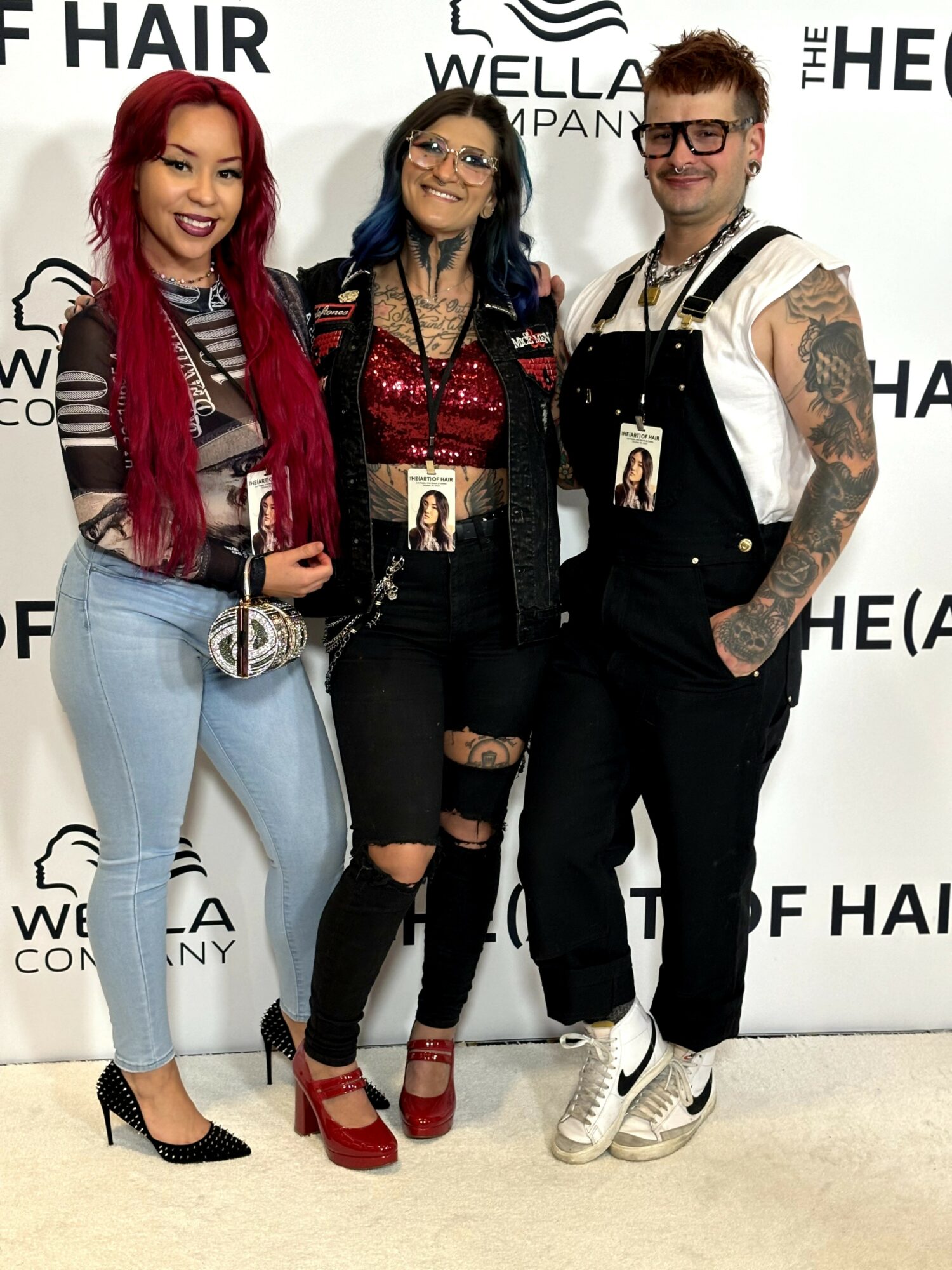Three people standing together in front of a backdrop with text, smiling, with one woman having red hair, another with glasses, and a man with tattoos.