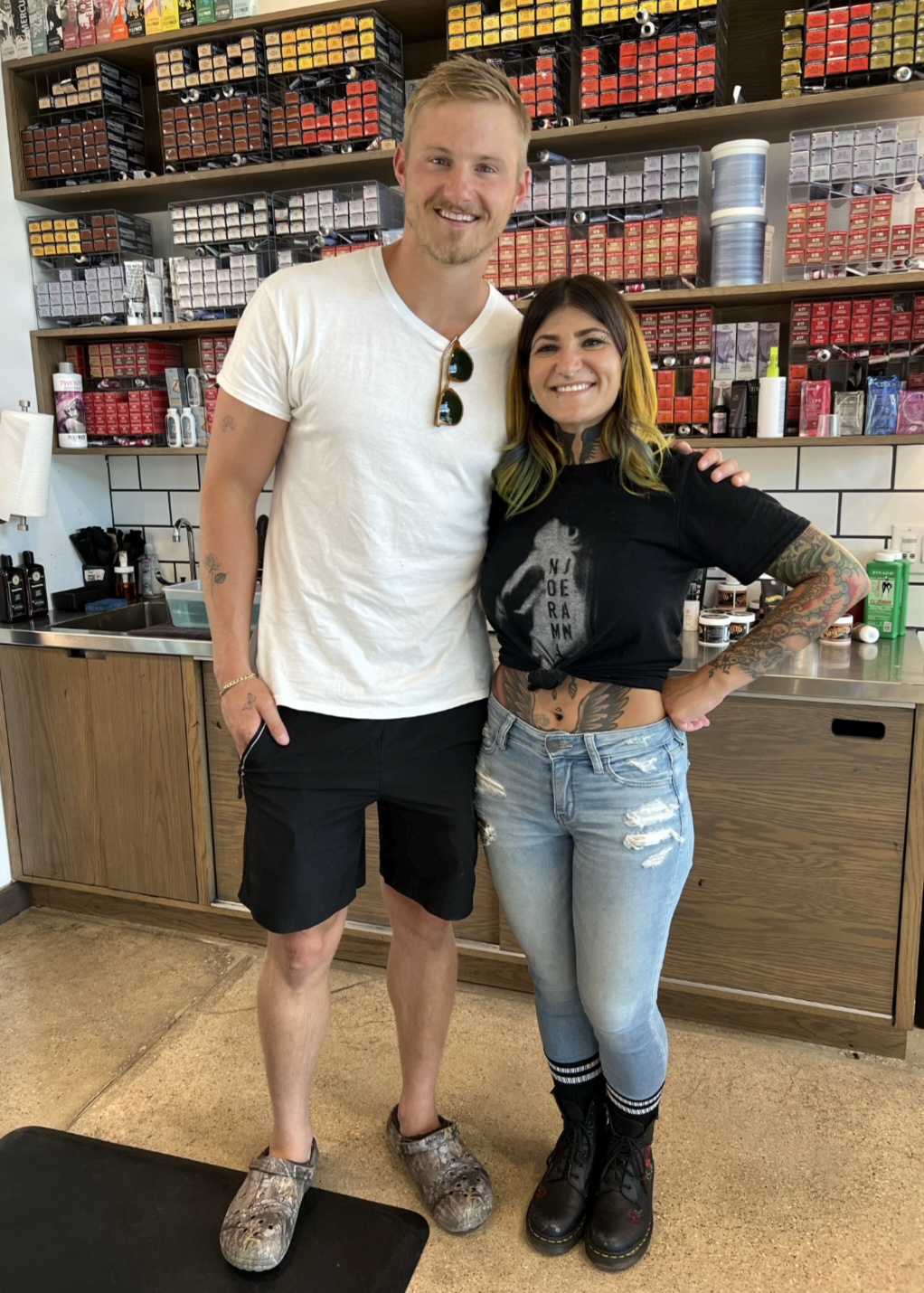 Two people standing in a store with shelves of colorful products behind them, smiling, one with arm around the other.