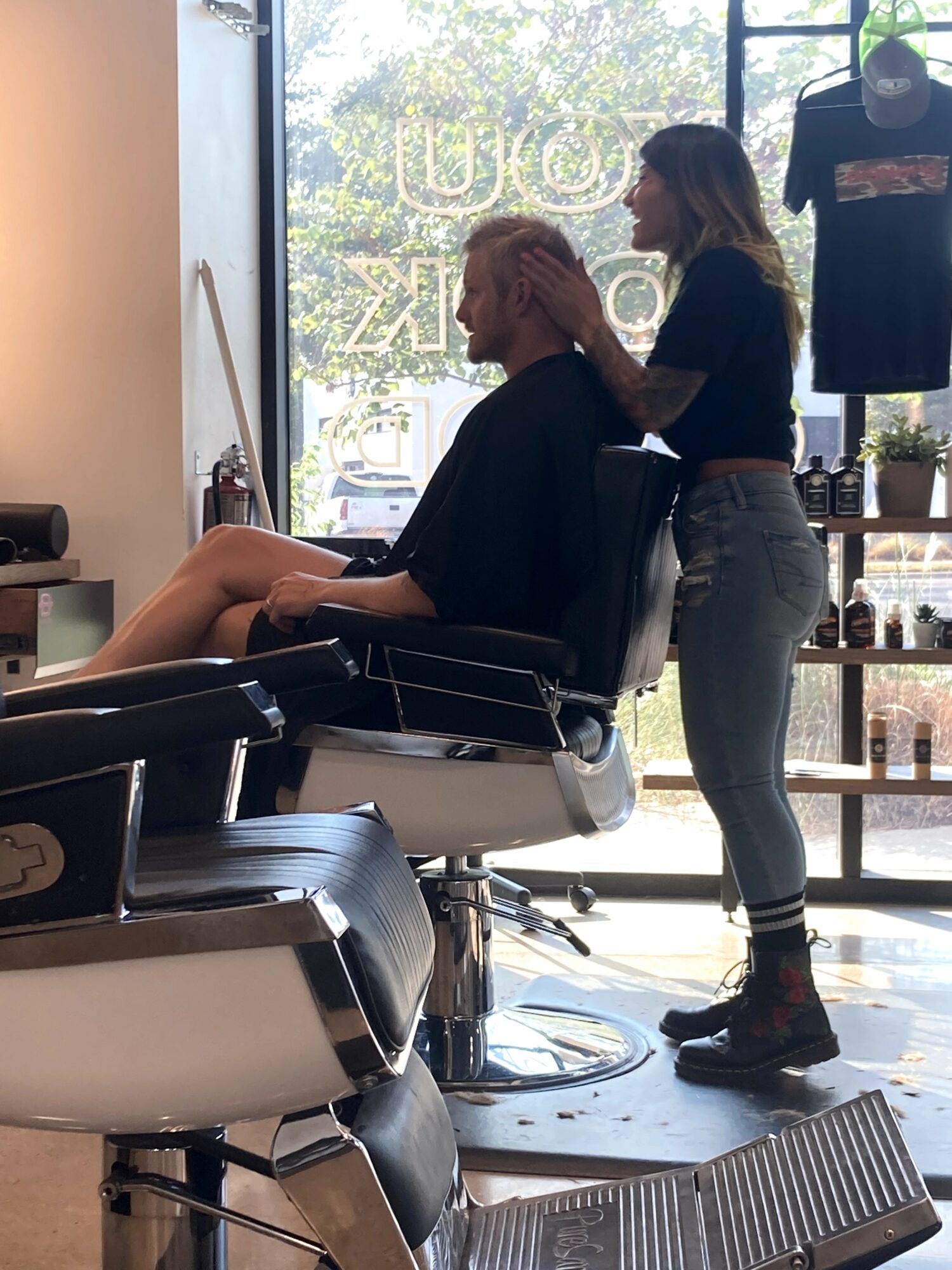 Person sitting in a salon chair with a stylist standing behind, inside a hair salon with large window and products on shelves.