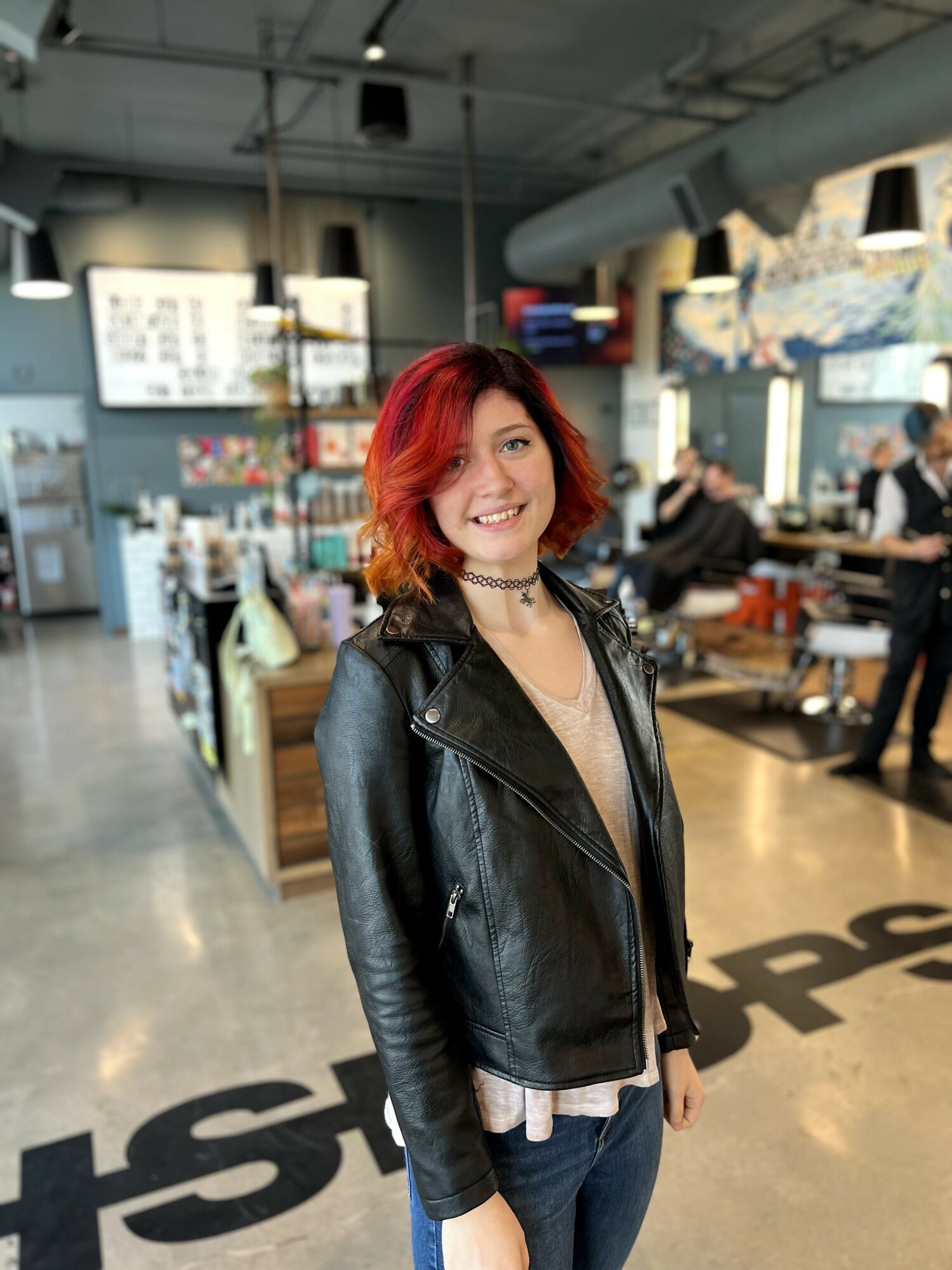 Young woman with red hair smiling in a cafe, wearing a black leather jacket and choker, with people and cafe decor in background.